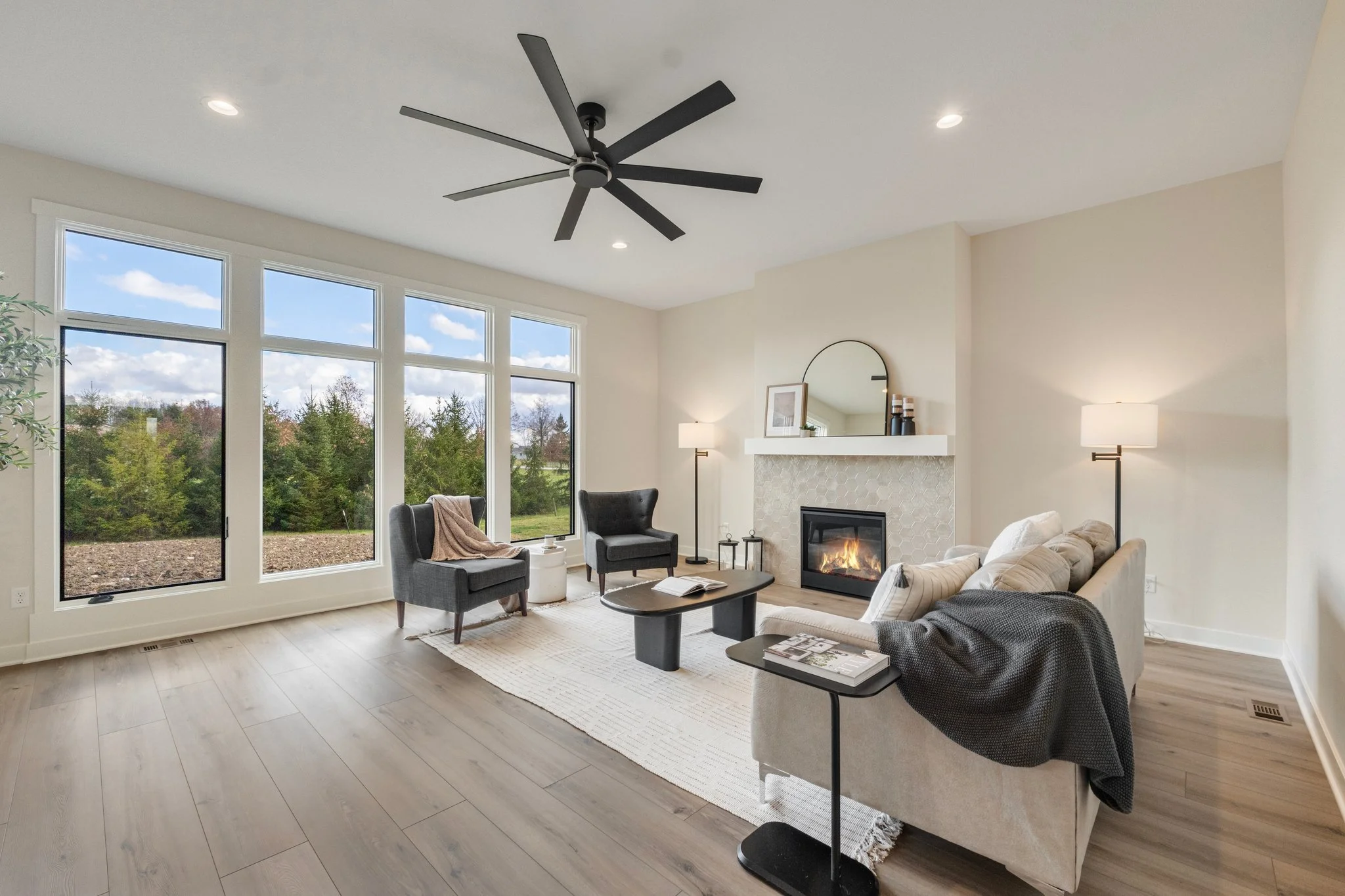 Modern living room with large windows, a fireplace, a beige sofa with pillows, two gray armchairs, a black coffee table, and floor lamps.