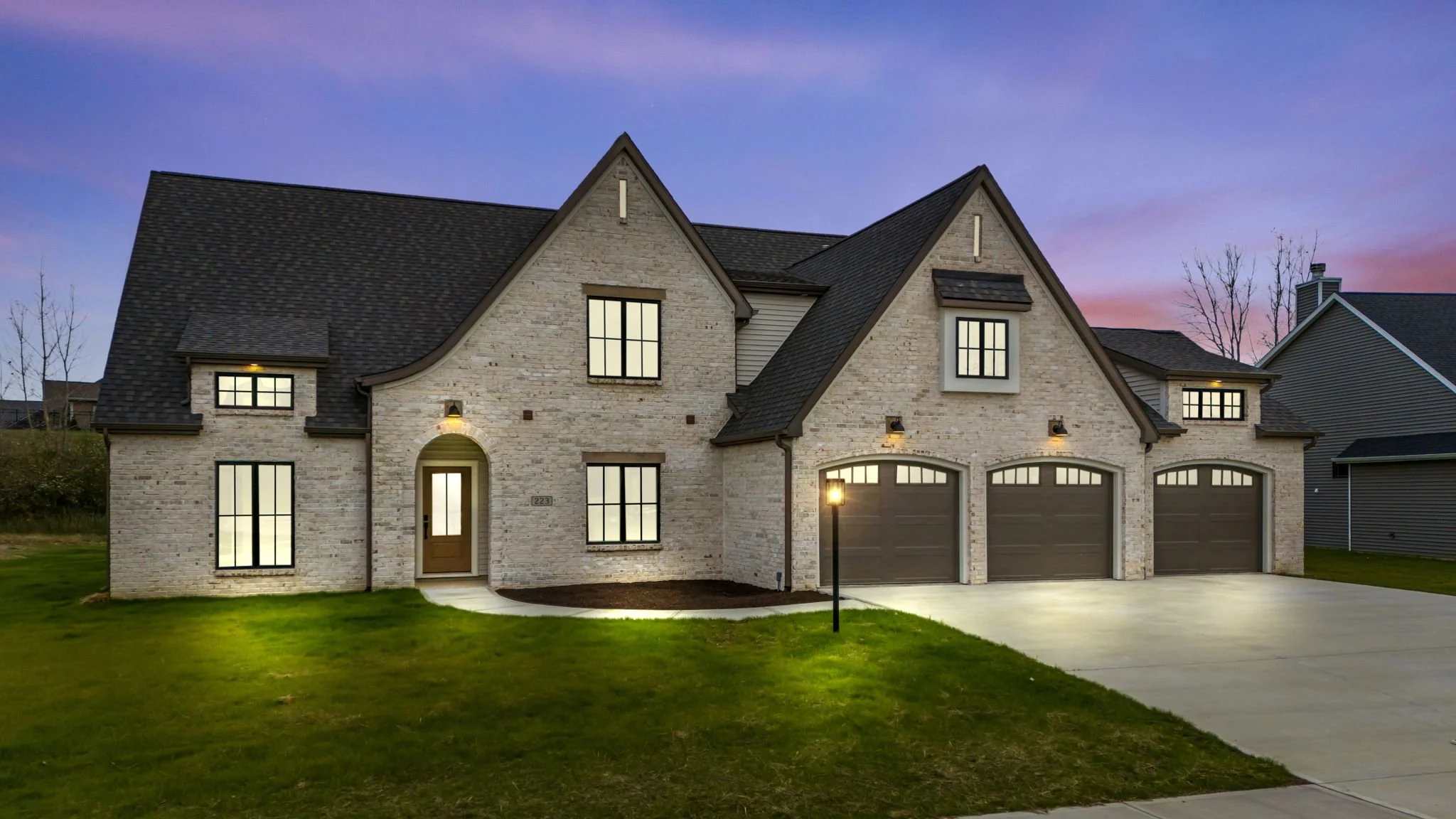 Modern two-story house with brick exterior, black shingle roof, and three-car garage, illuminated outside at dusk.