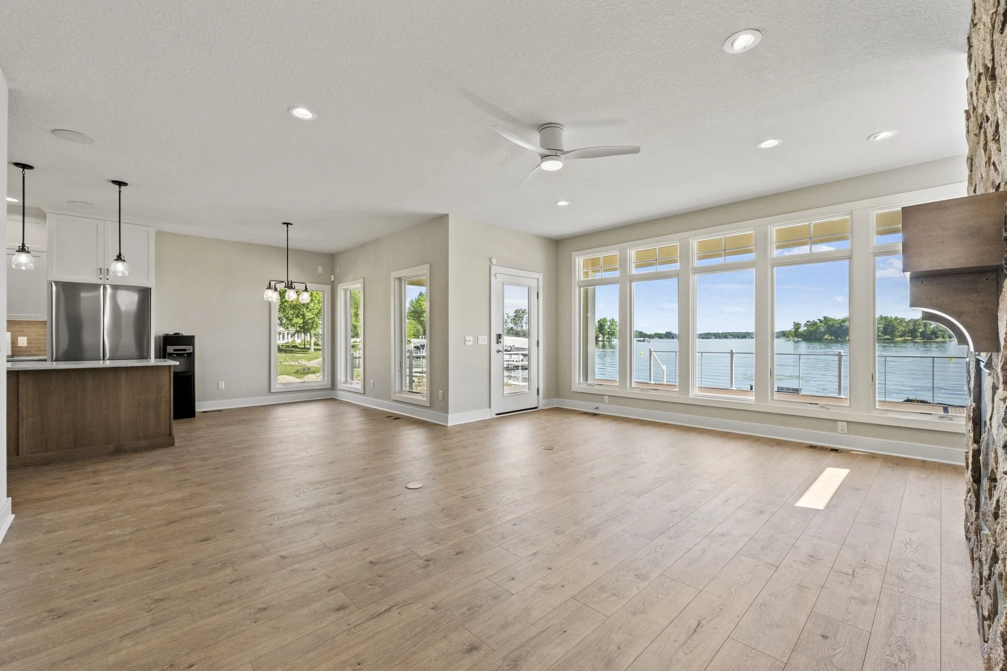 Empty living room with large windows overlooking a lake, wooden floors, modern ceiling fan, and a partial view of the kitchen.