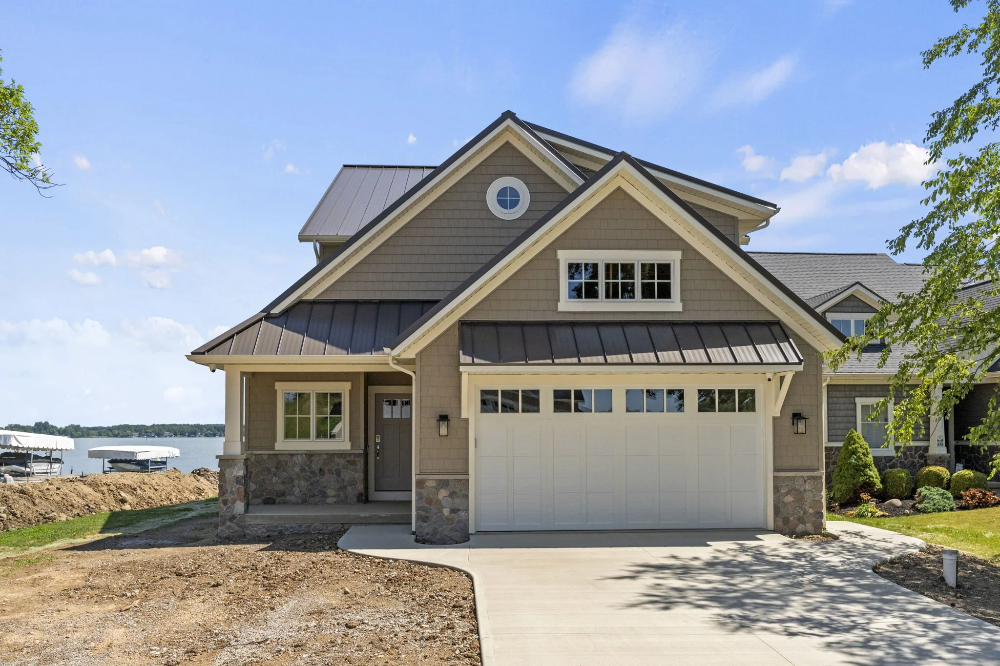 New house with a garage, stone accents, and a small front porch, situated near a body of water with boats and lush landscaping.