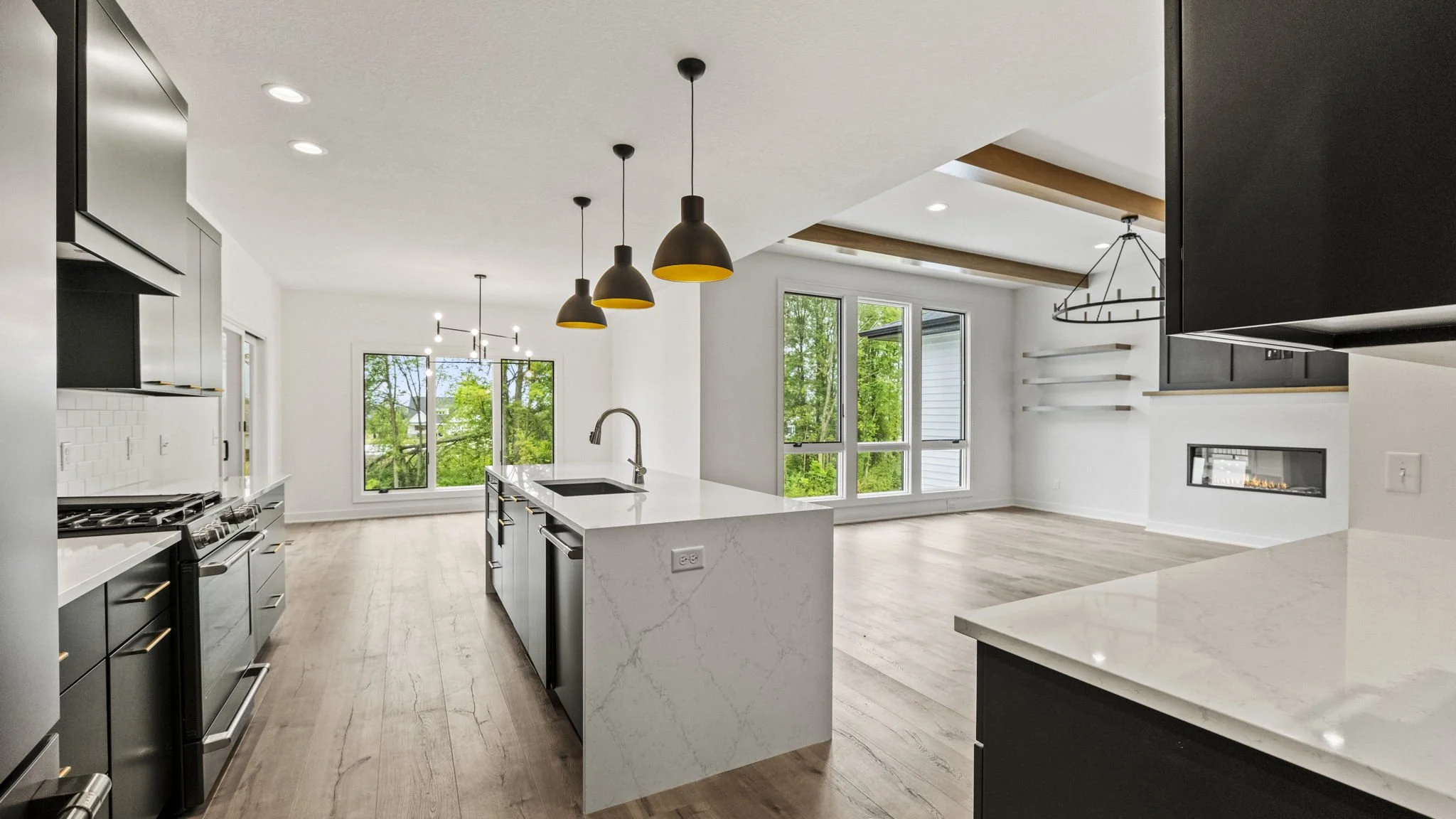 Modern open-plan kitchen and living room with large windows, white walls, black cabinetry, wood accents, and a fireplace.