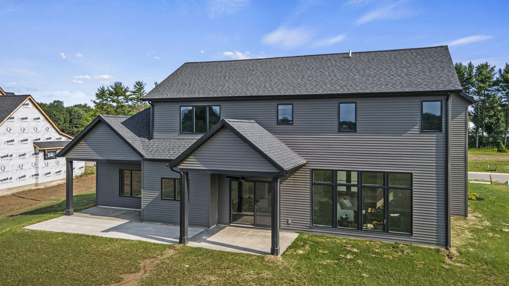 Newly constructed modern house with gray siding, large windows, and a small covered patio, situated on a grassy lot under a blue sky.