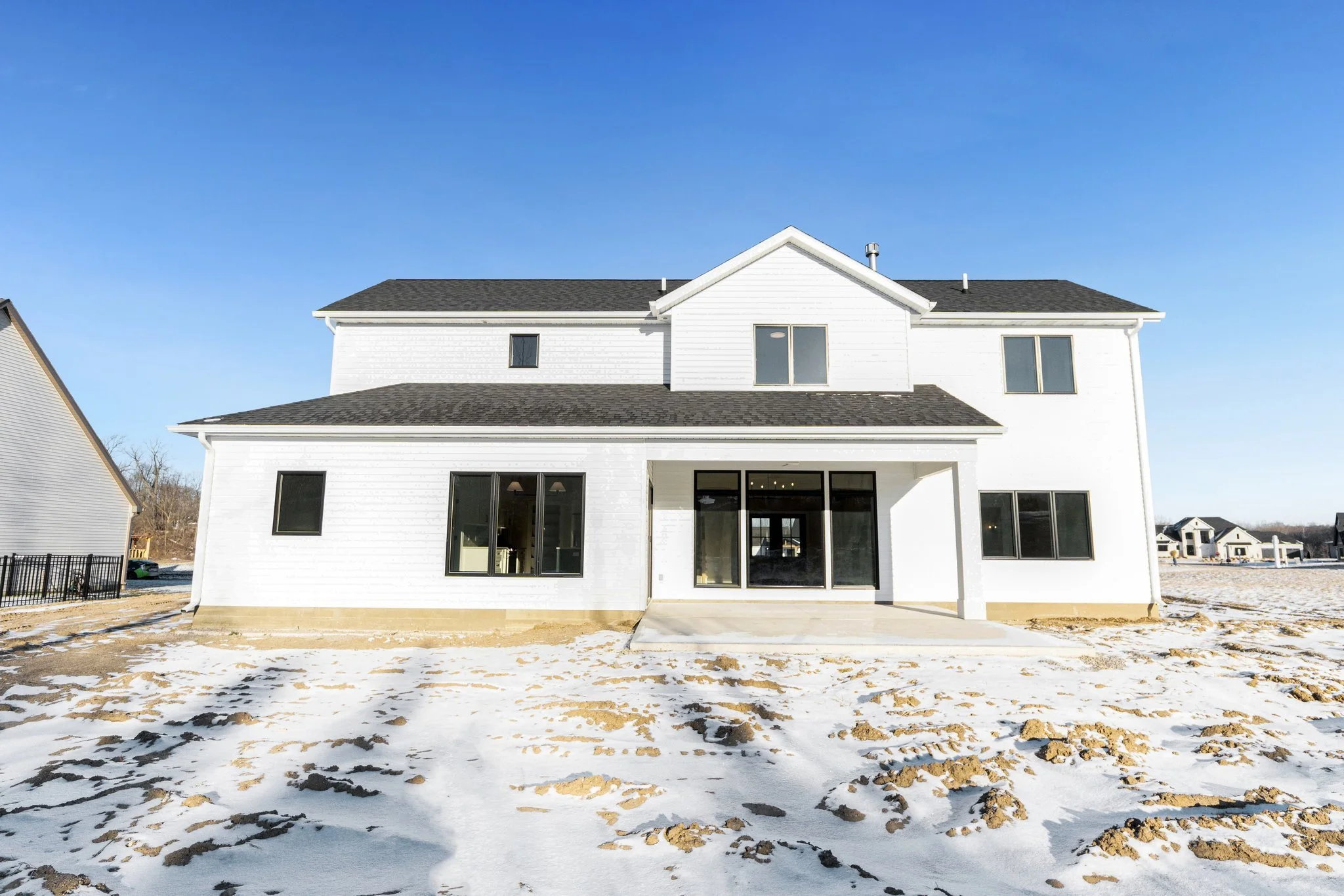 Newly constructed two-story white house with black roofing, large windows, and a concrete patio, situated in a snowy landscape with clear blue sky.