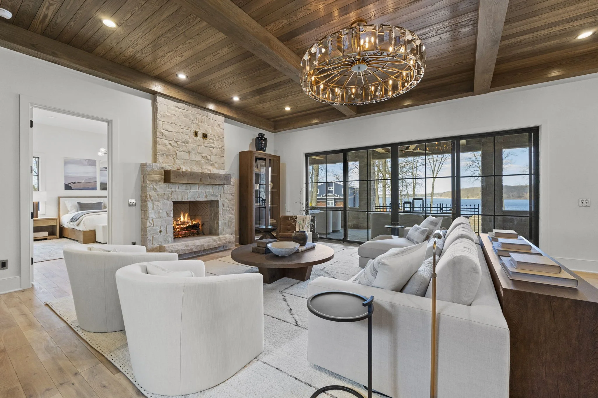 Living room with white sofa and armchairs, stone fireplace with fire, large sliding glass doors overlooking water, wooden ceiling with chandelier, bookshelf with books.
