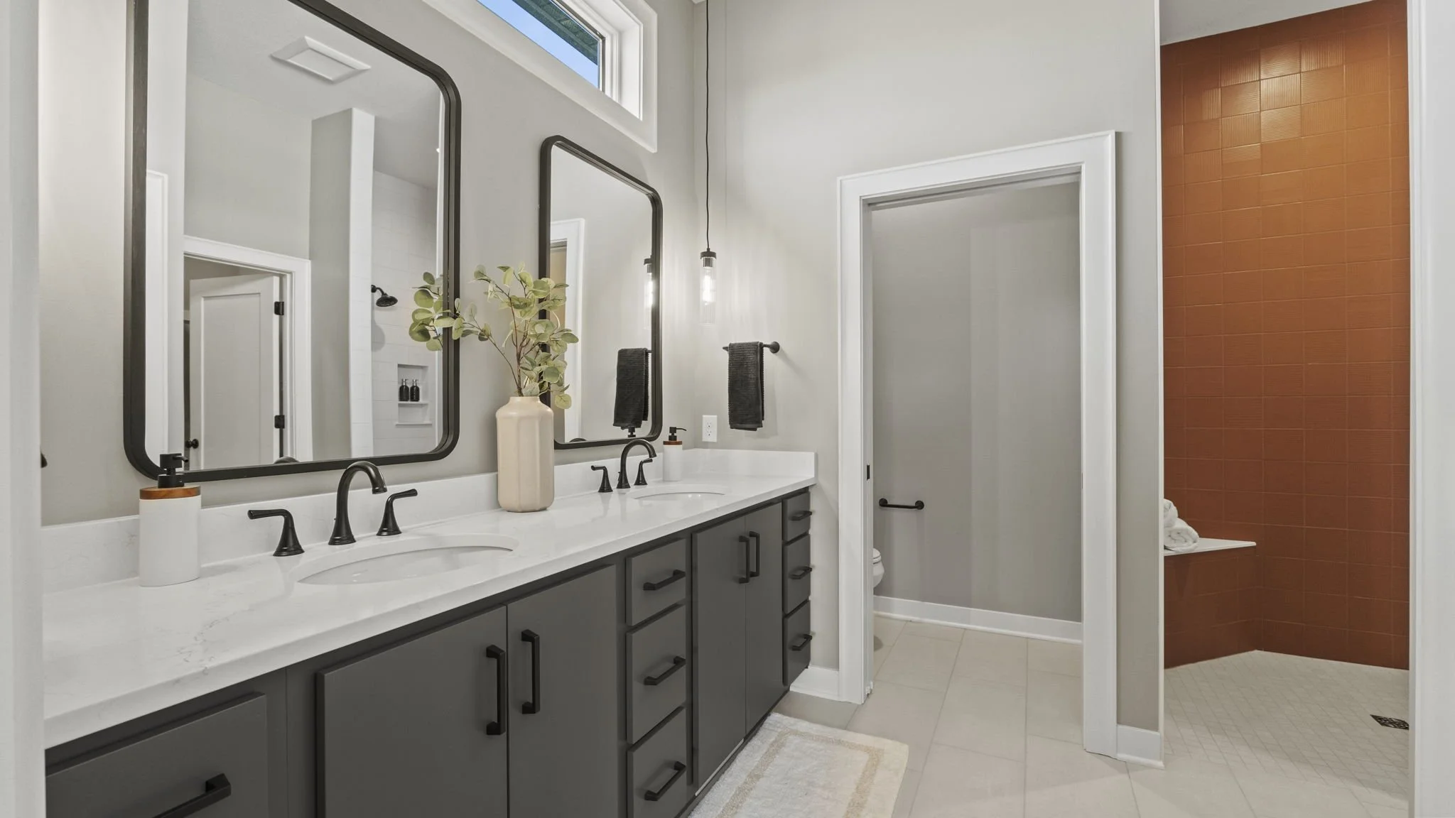 Modern bathroom featuring a double vanity with gray cabinets, black hardware, white marble countertops, two large rectangular mirrors, and pendant lights. There is a decorative vase with greenery, and a towel holder with dark towels. The bathroom inc