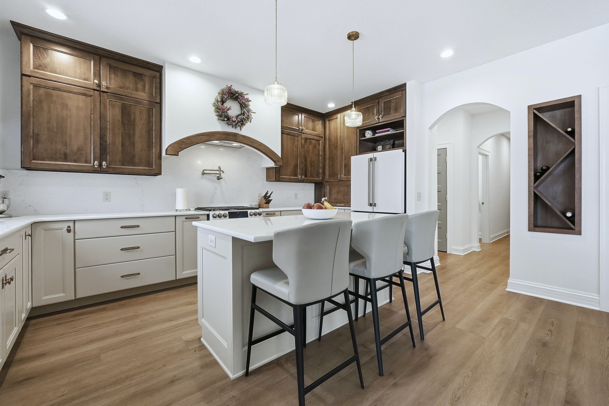 Modern kitchen with white and wood cabinets, kitchen island with three chairs, wooden flooring, pendant lights, decorated with a wreath and bowl of fruit.