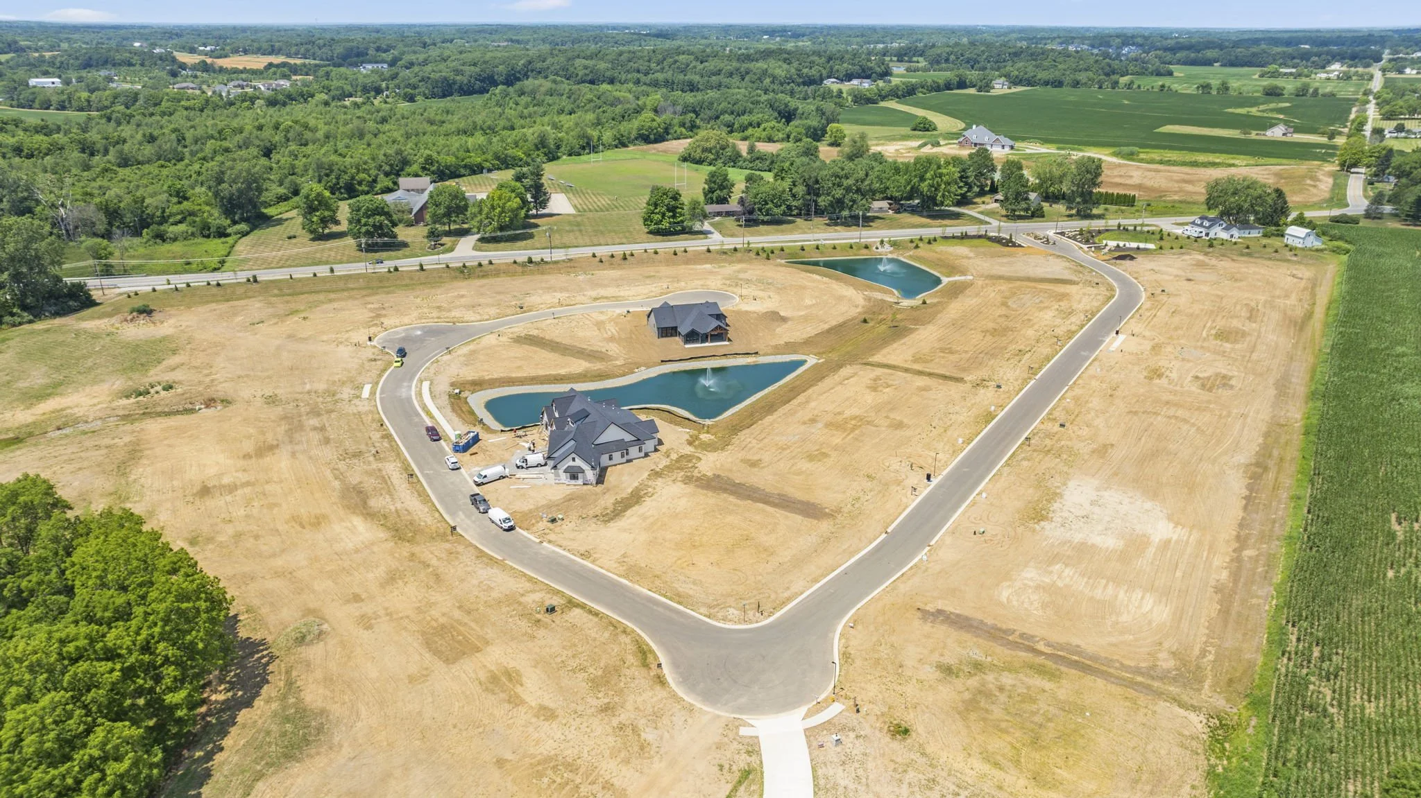 Aerial view of a new residential development with a house, three ponds with fountains, and an asphalt road layout in a rural area with fields and trees.