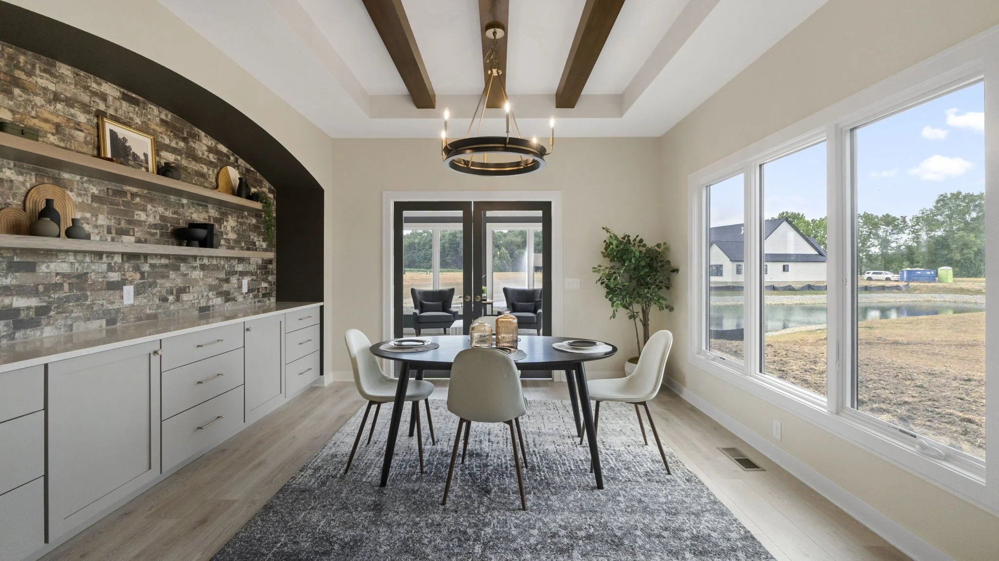 Modern dining room with round black table and six white chairs, large windows overlooking a backyard, and double glass doors leading to an outdoor seating area with two black chairs, a chandelier hanging from ceiling with exposed wooden beams, and a 