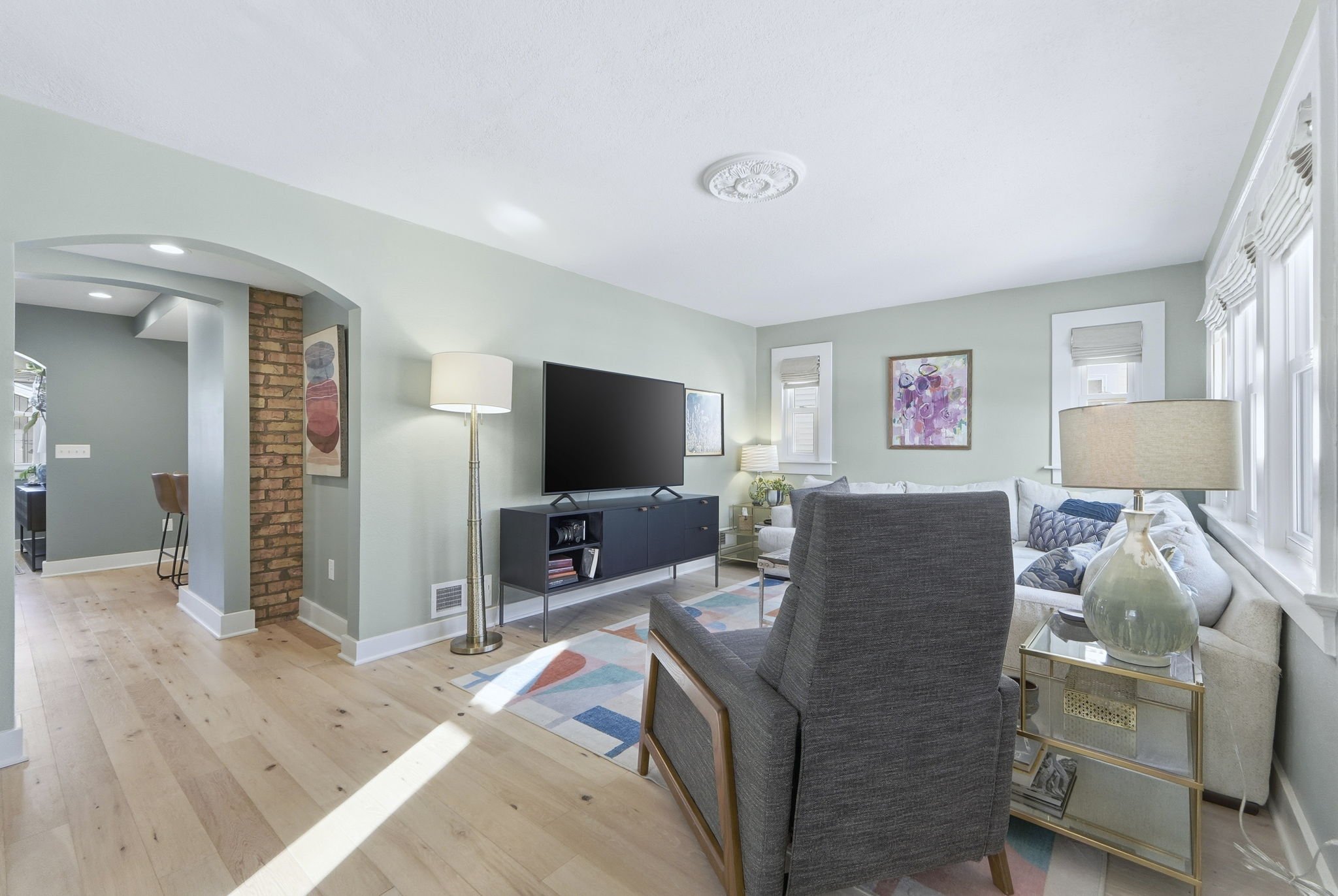 Living room with pastel green walls, a large white sectional sofa with colorful pillows, a dark gray armchair, a glass side table with a table lamp, a TV on a black cabinet, and abstract artwork on the walls. Light wooden flooring and multiple window