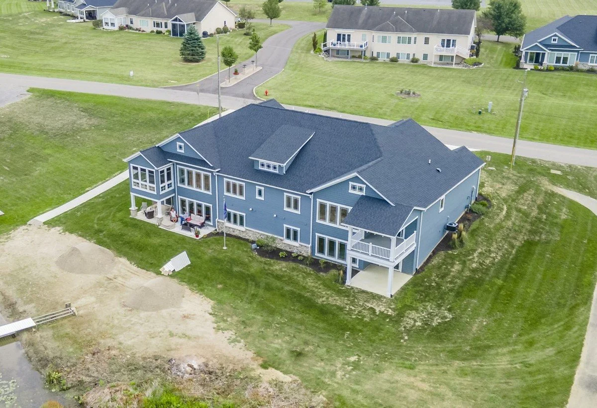 An aerial view of a large blue house with a dark roof, multiple decks, and landscaped yard, situated in a suburban neighborhood with other houses, roads, and green lawns.