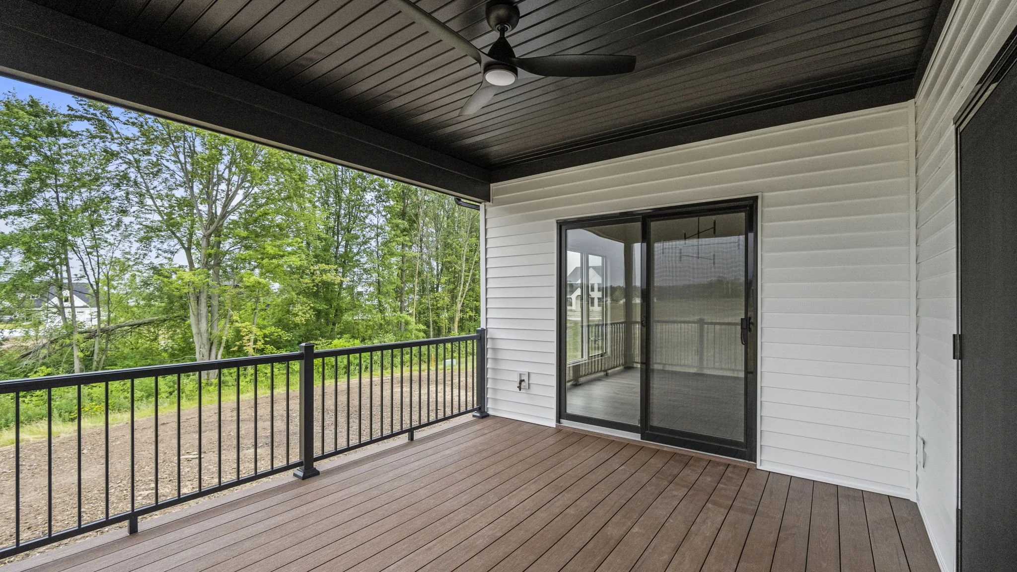 Empty outdoor balcony with brown wood flooring, black metal railing, white siding walls, a black ceiling fan, and glass sliding door, overlooking green trees and a partly cloudy sky.