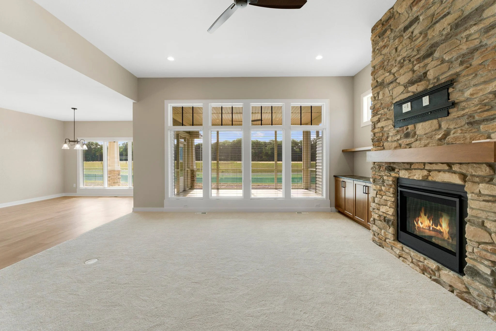 Empty living room with large windows, a stone fireplace with a wooden mantel, and a ceiling fan, overlooking a backyard with a porch.