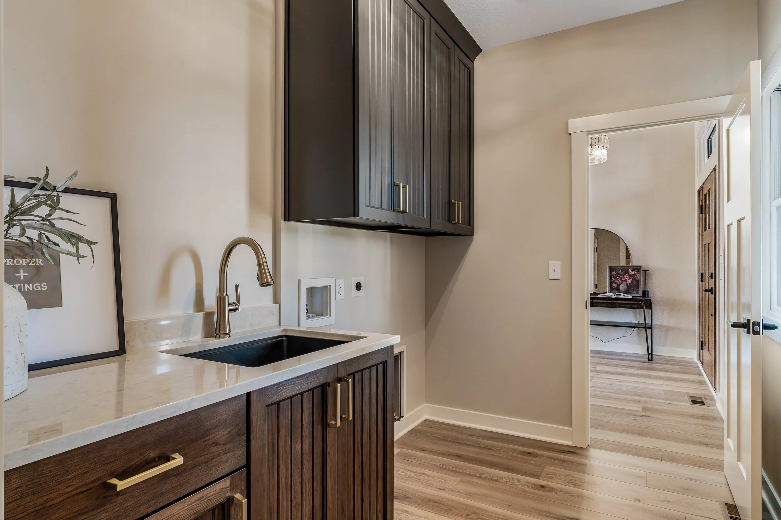 Interior view of a kitchen with dark wooden cabinets, a beige countertop with a black sink, a brass faucet, and a framed quote on the counter. A doorway leads to a room with a small desk and chair, wood flooring, and neutral-colored walls.