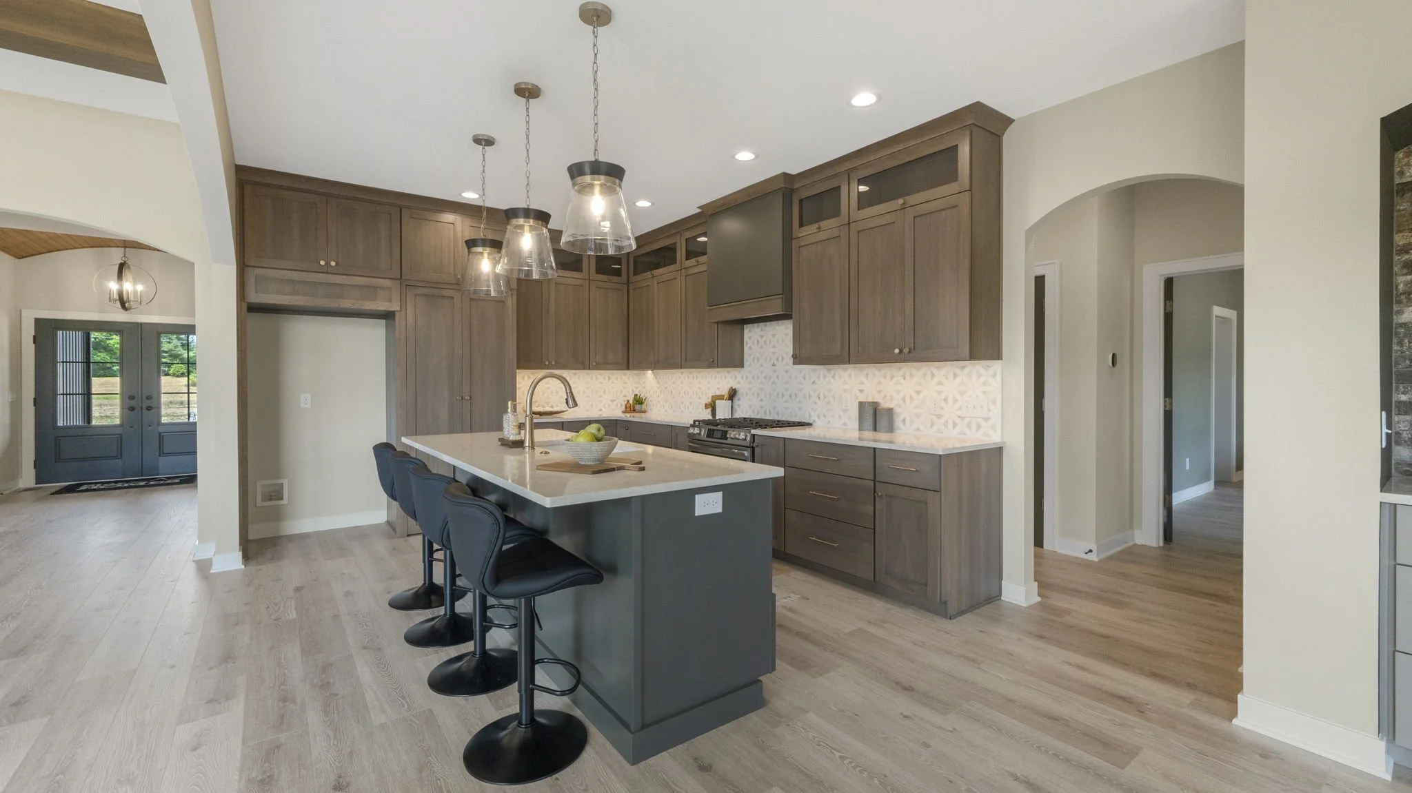 Modern kitchen with wooden cabinetry, white countertops, and a kitchen island with four black barstools. Pendant lights hang above the island. Light-colored hardwood floors and an open layout with double doors leading outside.