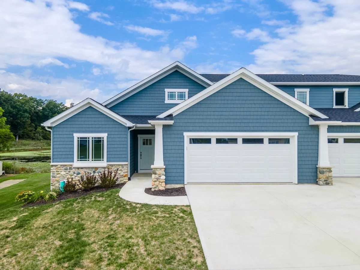 Front view of a modern blue house with a white garage door, stone accents, and a small porch under a partly cloudy sky.