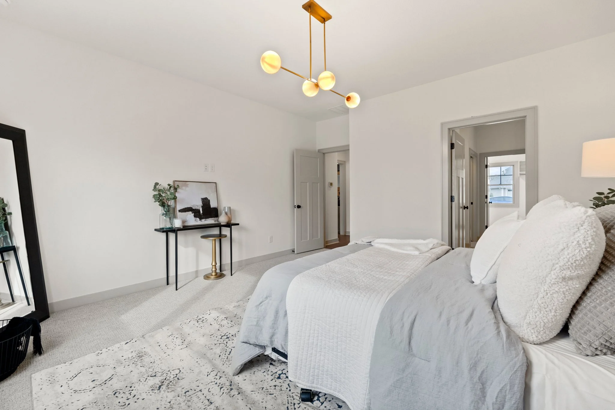 A bedroom with white walls and a beige carpeted floor. There is a bed with white bedding and pillows on the right side. A black framed mirror and a small black table with decorative items including a framed artwork, a vase with green foliage, and a l