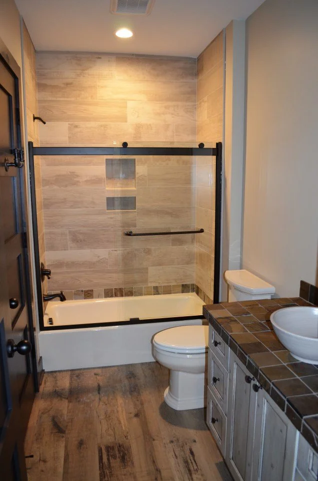 Bathroom with a bathtub and shower, toilet, and a vanity with a sink. The shower has glass doors and a grab bar, with wood-look tiles on the wall and a tiled floor. The vanity has a tiled countertop and wooden cabinets.