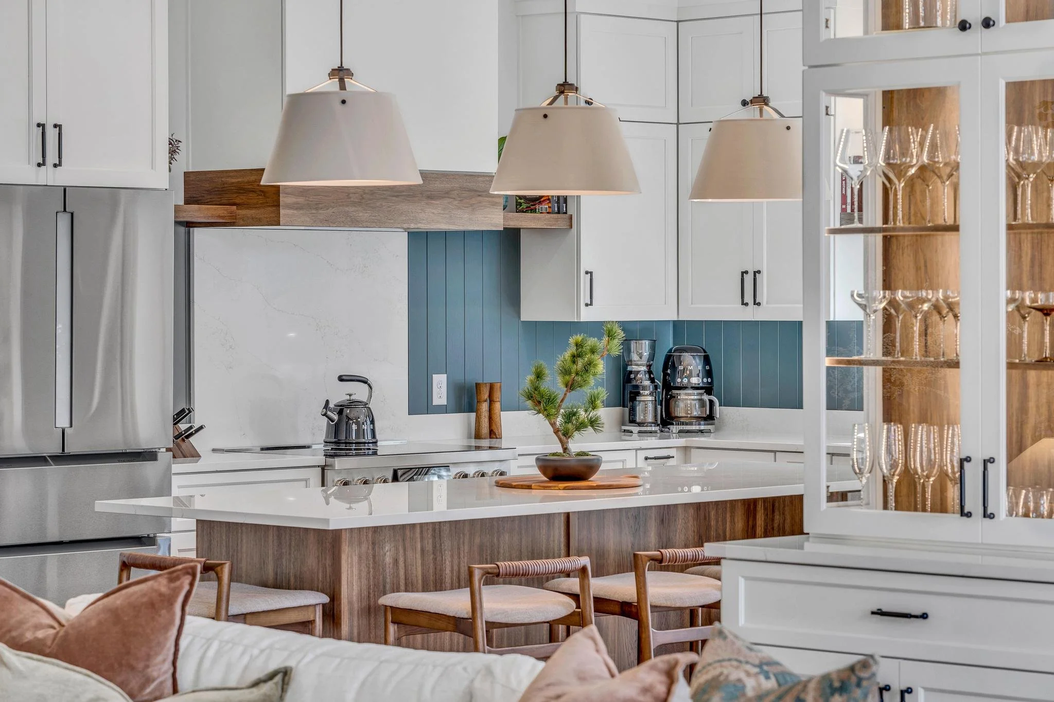 Modern kitchen with white cabinets, blue backsplash, wooden accents, pendant lighting, and a small potted plant on the island.