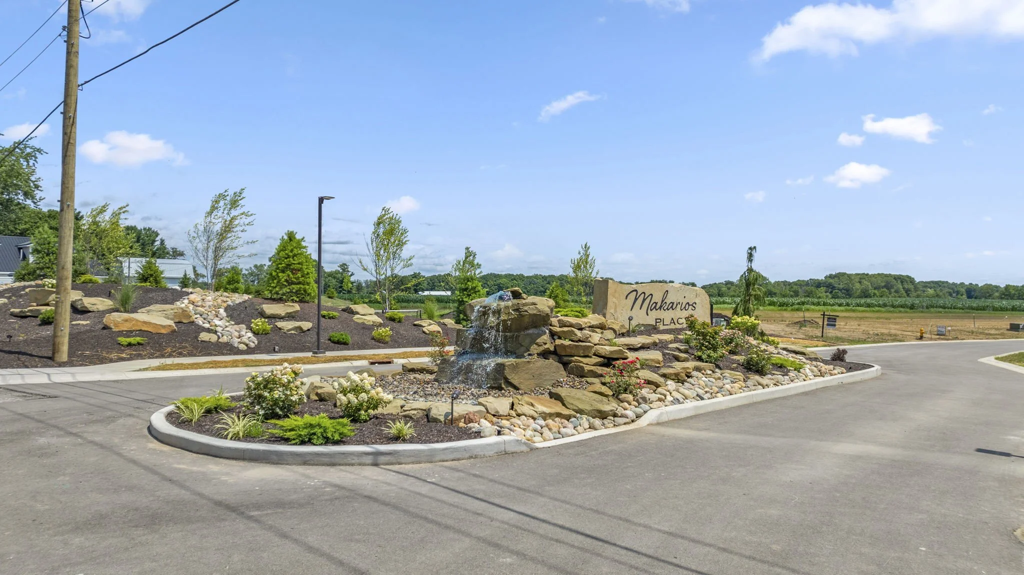 Entrance sign for Makarios Place with a rock fountain and landscaped garden bed, under a partly cloudy sky.