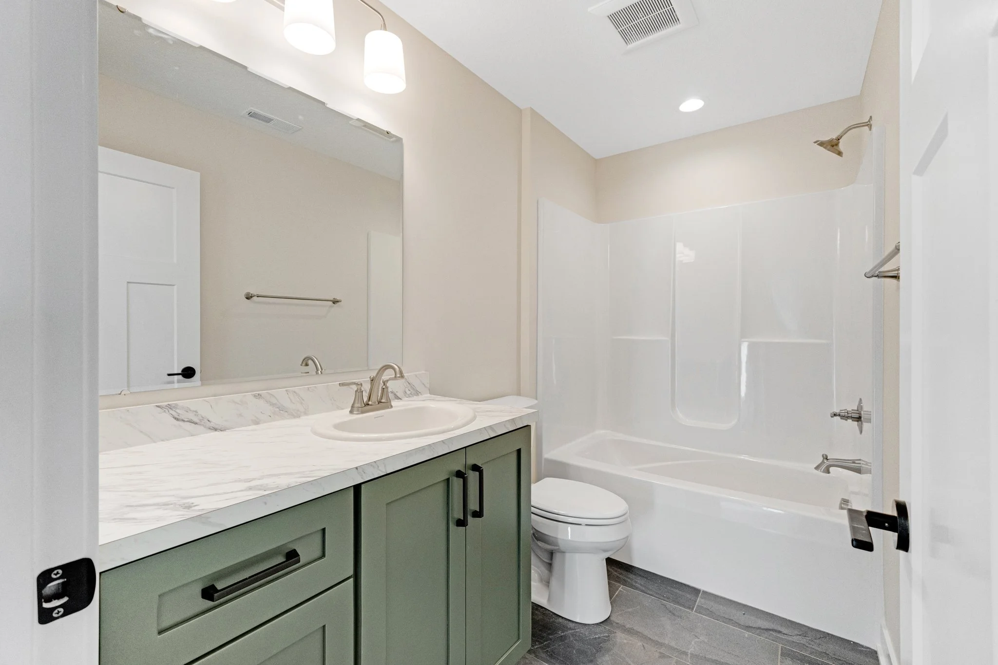 Modern bathroom with green vanity, marble countertop, large mirror, white bathtub with shower, toilet, and grey floor tiles.