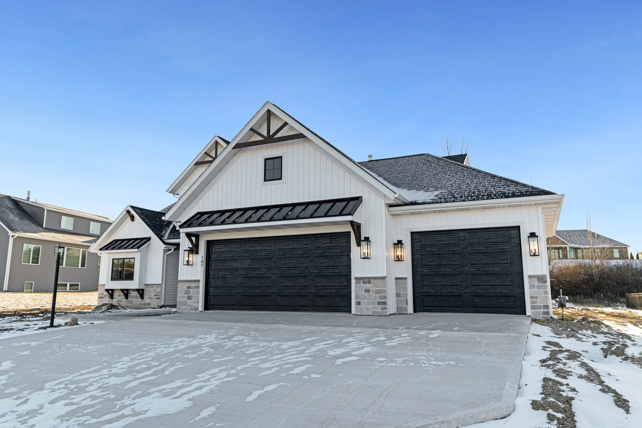 A modern two-story house with a white exterior, black garage doors, and black trim, situated in a snowy landscape under a clear blue sky.
