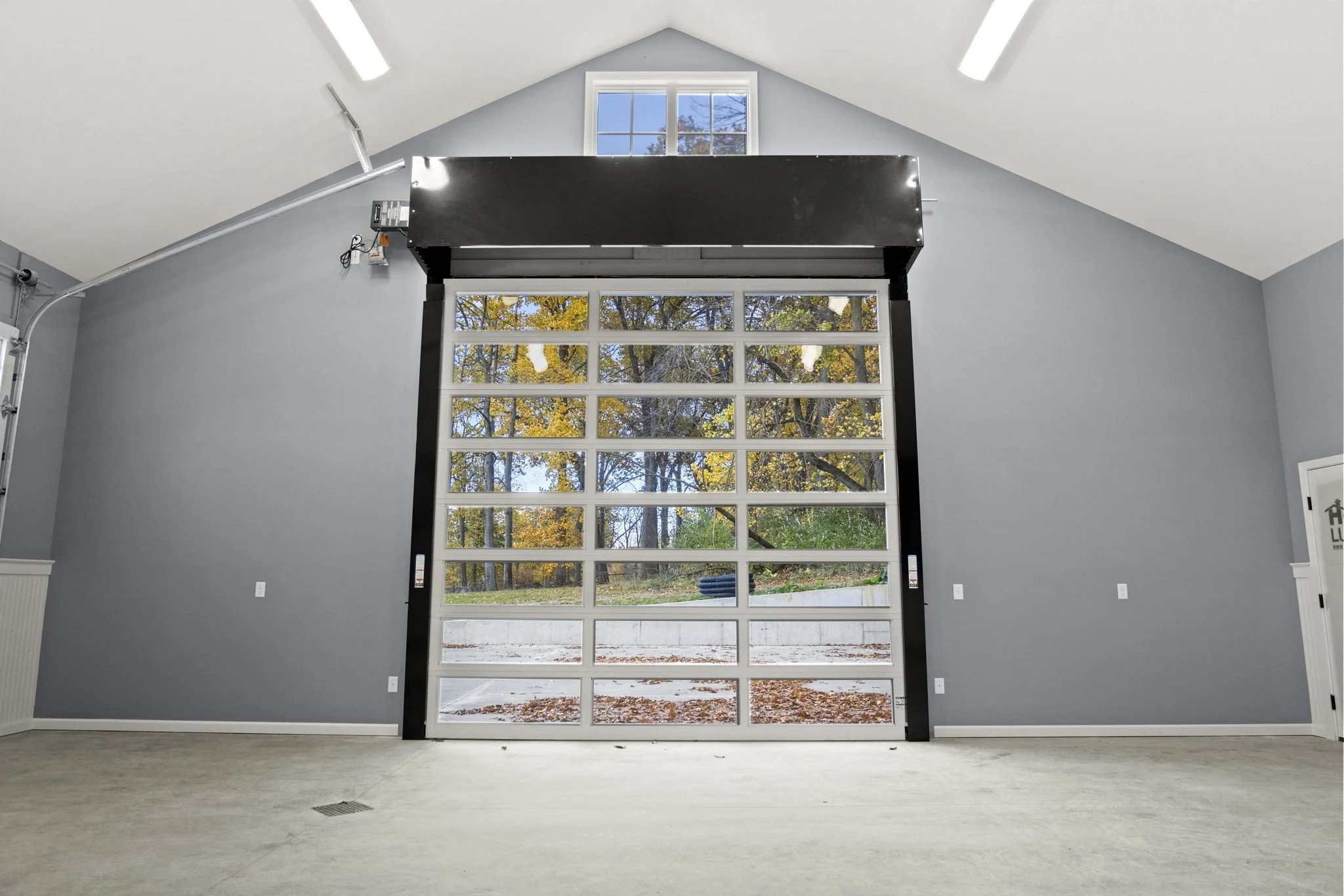 Empty garage with a large glass-paneled door, painted gray walls, vaulted ceiling, and window showing trees outside.