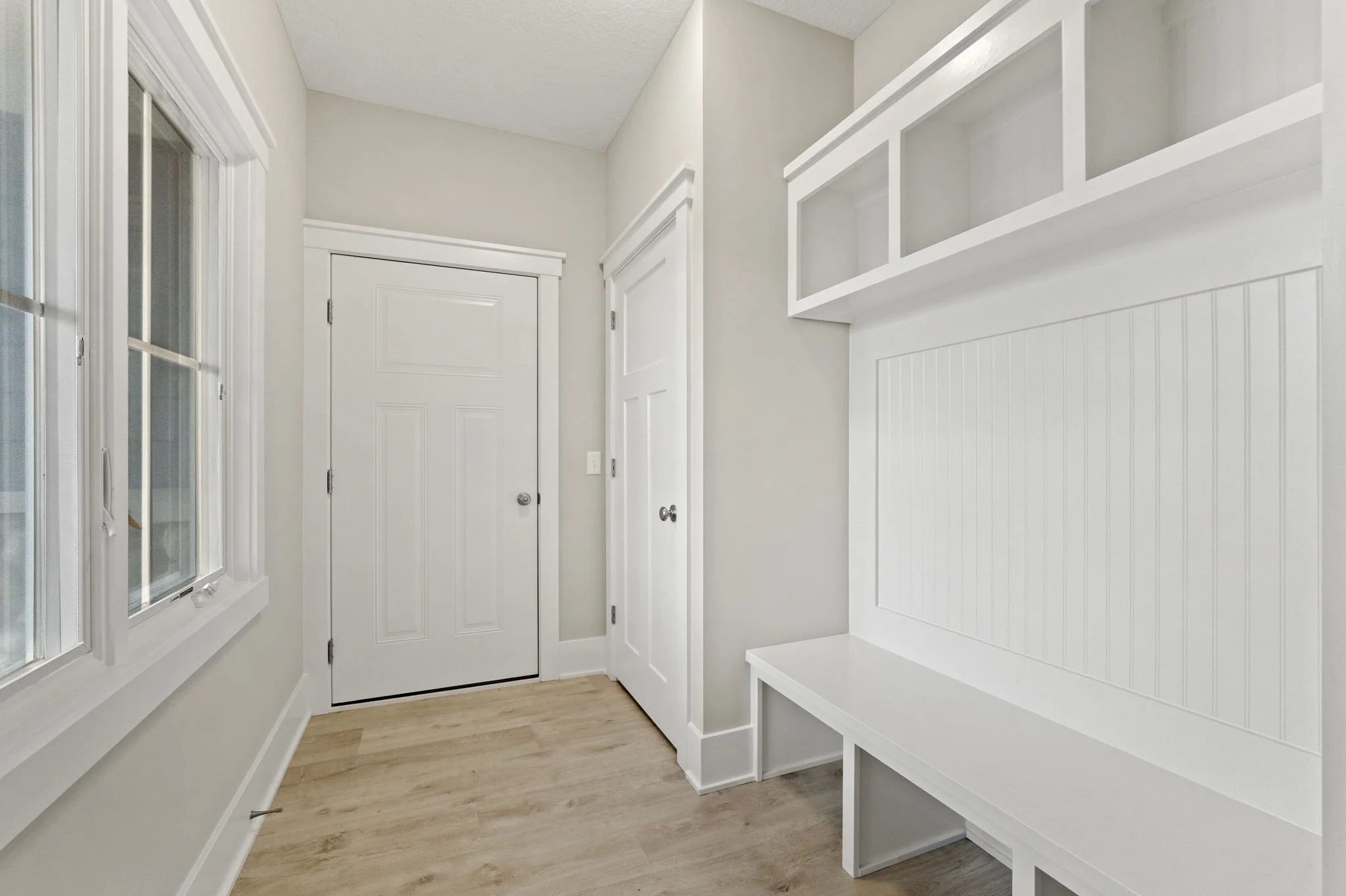 A bright, empty room with light wood flooring, white walls, a window on the left, and a white built-in bench with storage above on the right.