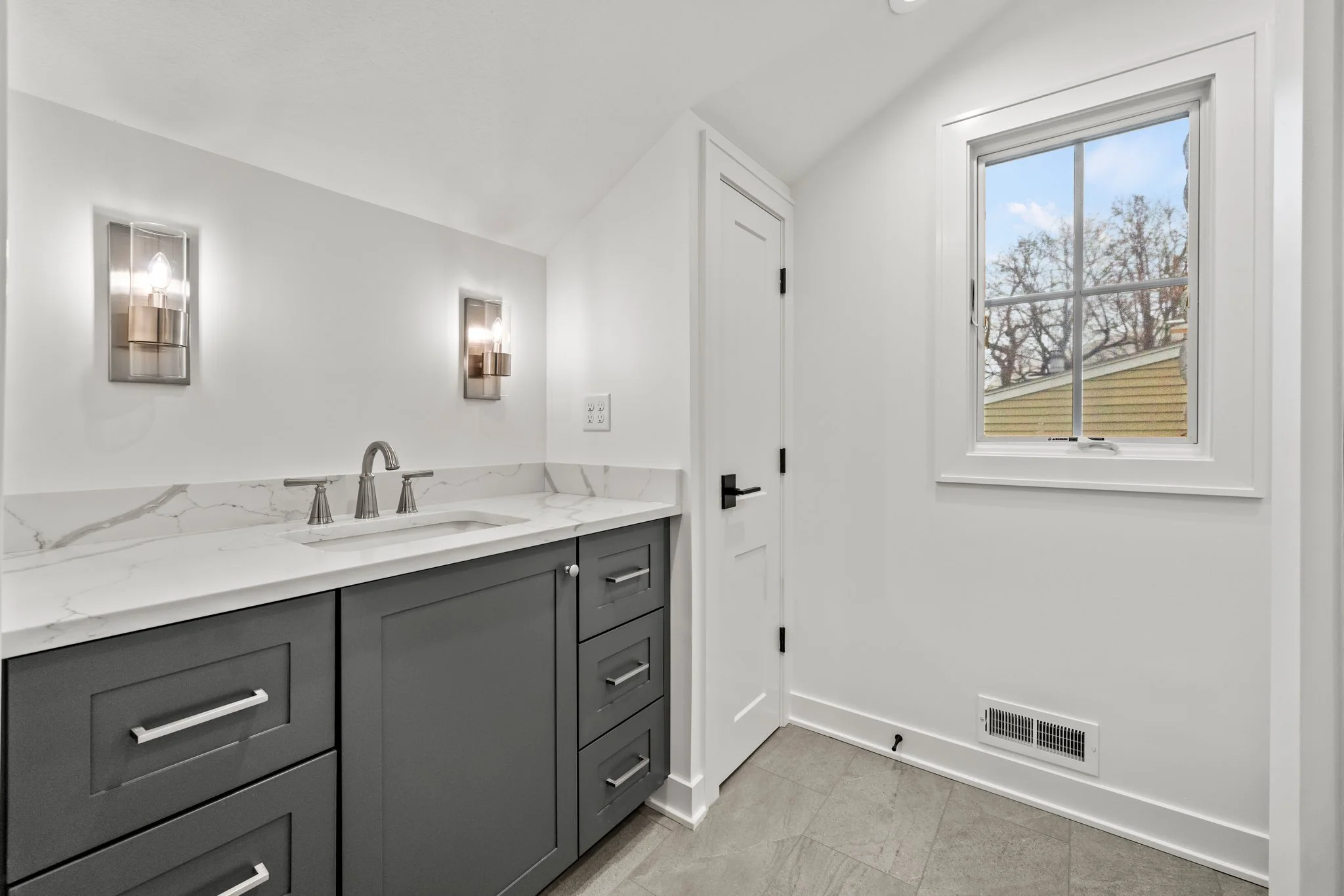 A modern laundry or utility room with a grey cabinet, marble countertop, wall-mounted light fixtures, a window with a view of trees and a yellow house, and a door