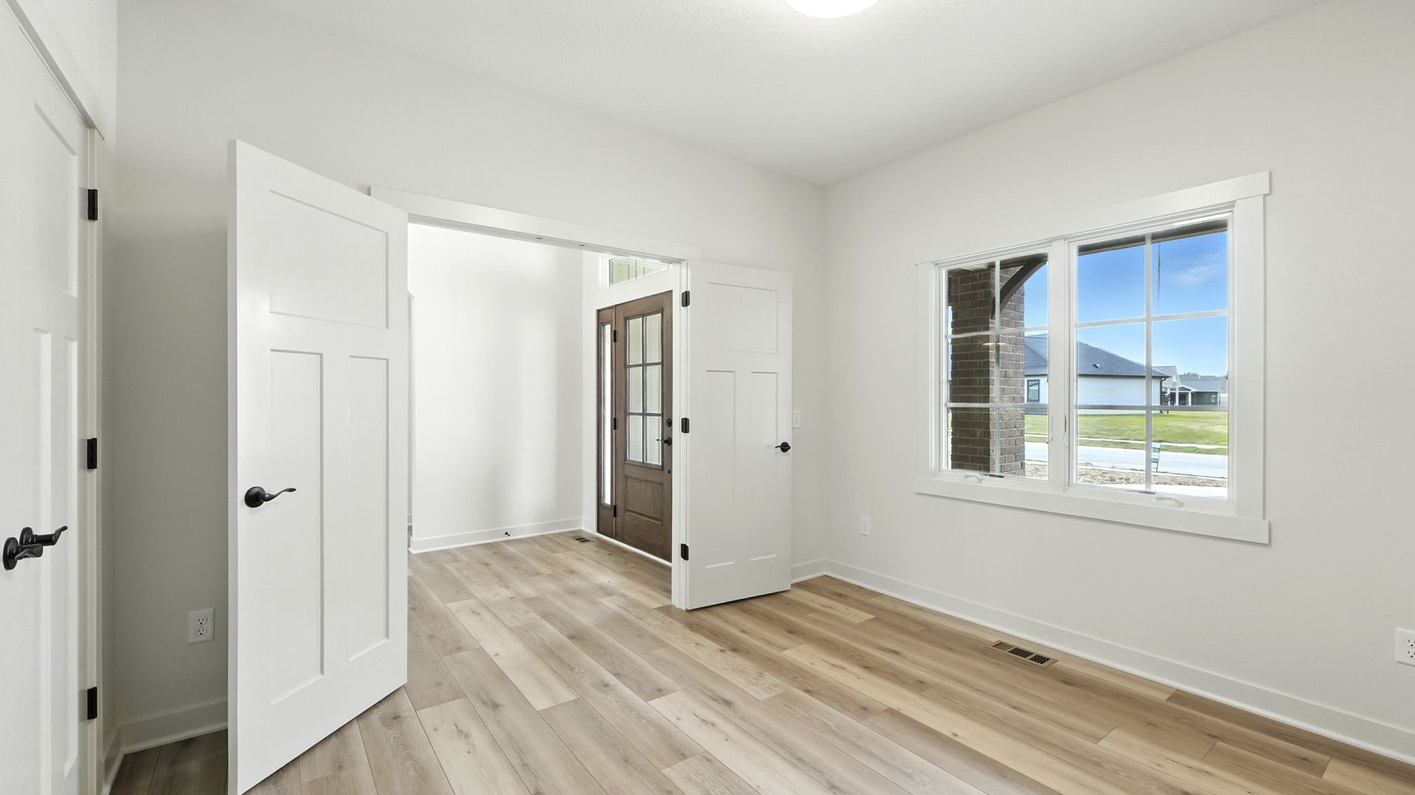 Empty room with wood flooring, white walls, a large window, and an open door leading outside. The room has a clean, modern look with minimal decor.