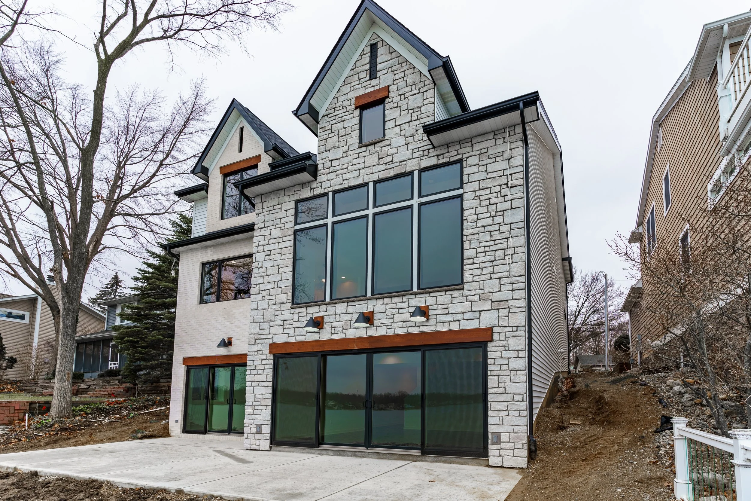 Modern three-story house with stone and siding exterior, large windows, and a paved driveway, surrounded by barren trees.