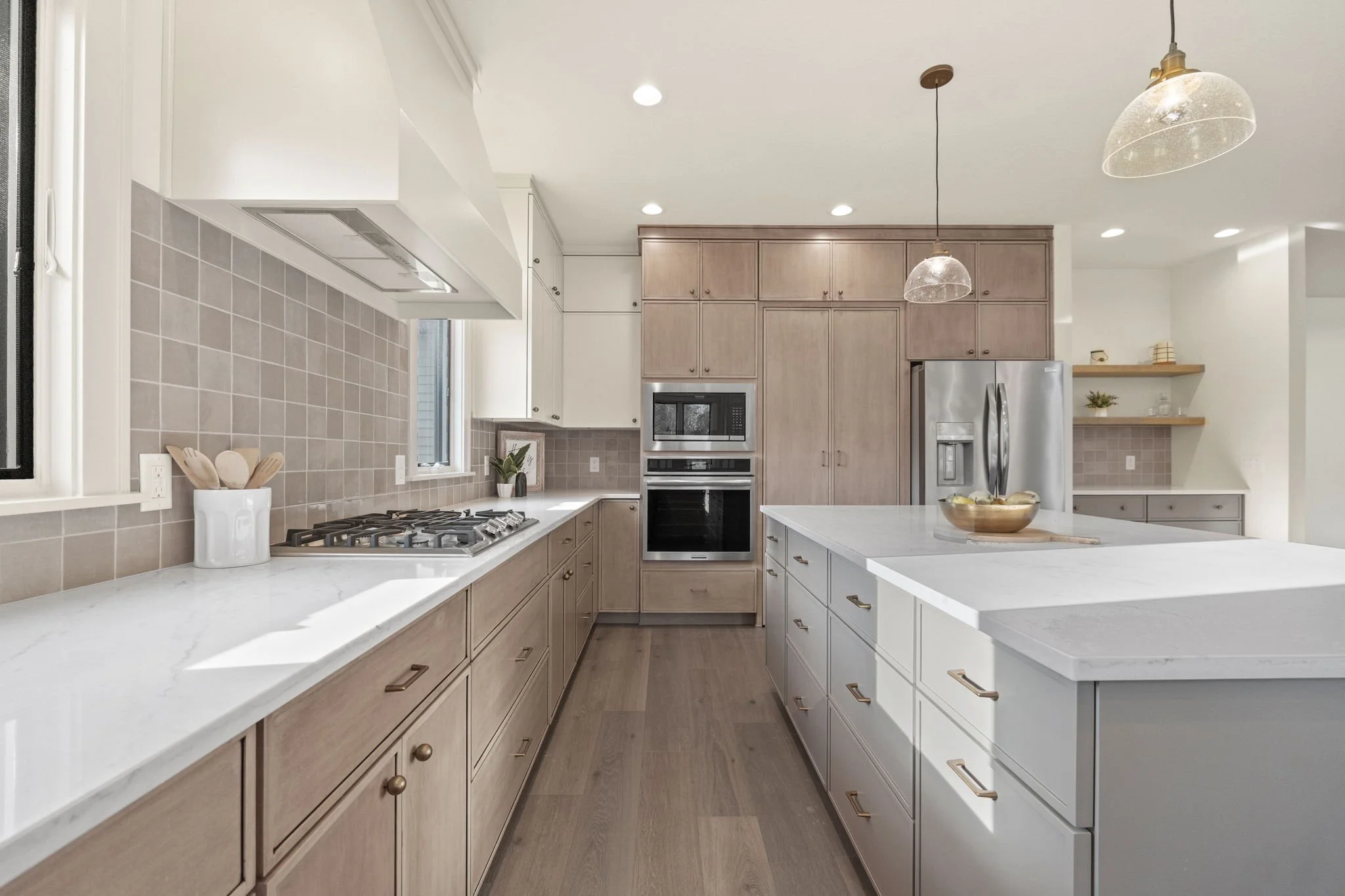 Modern kitchen with white and light wood cabinetry, a large island with white countertops, stainless steel refrigerator, built-in microwave and oven, beige tile backsplash, and wooden floating shelves.