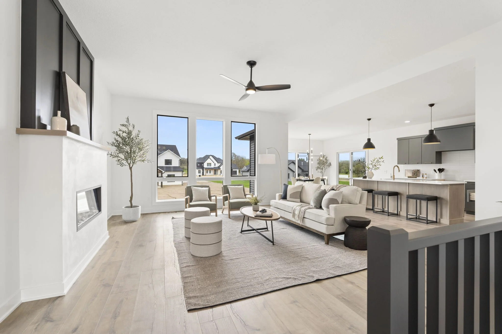 Bright modern living room with large windows, white walls, light wood floors, beige seating, black and gray accents, and an open kitchen with bar stools.