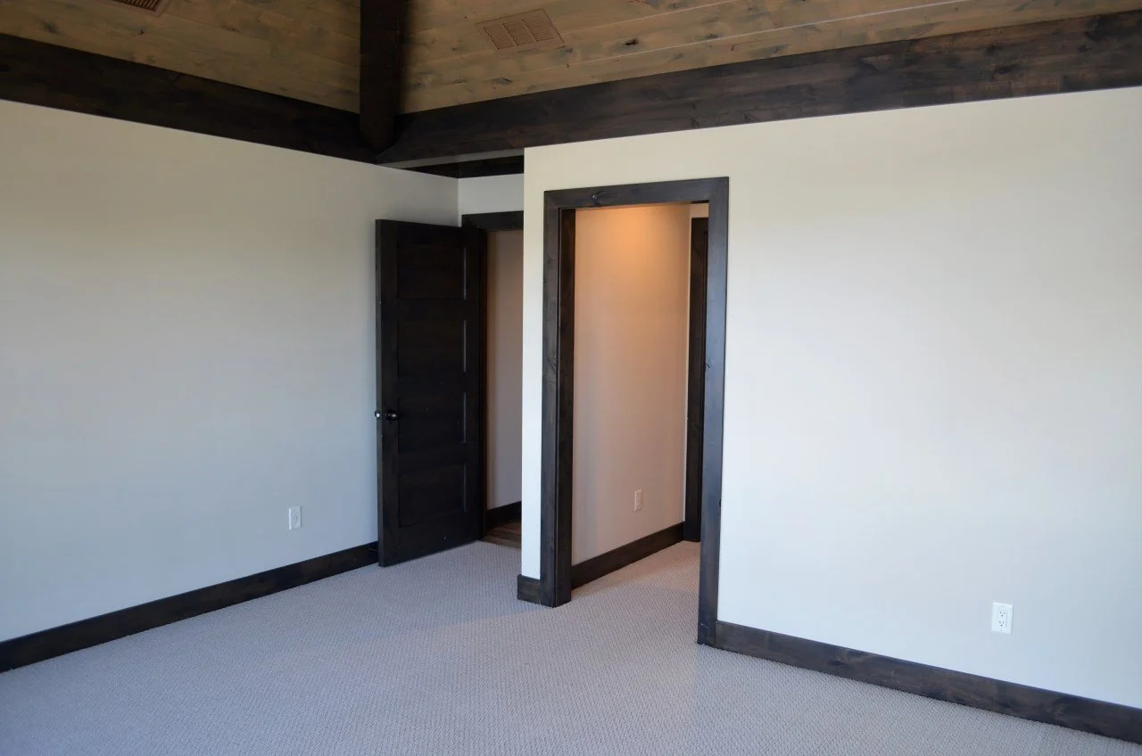 An empty room with beige walls, dark wood trim and door frames, carpeted floor, and a ceiling with dark wood paneling. There are two doorways, one with a dark wood door and the other open to a small hallway or closet space.