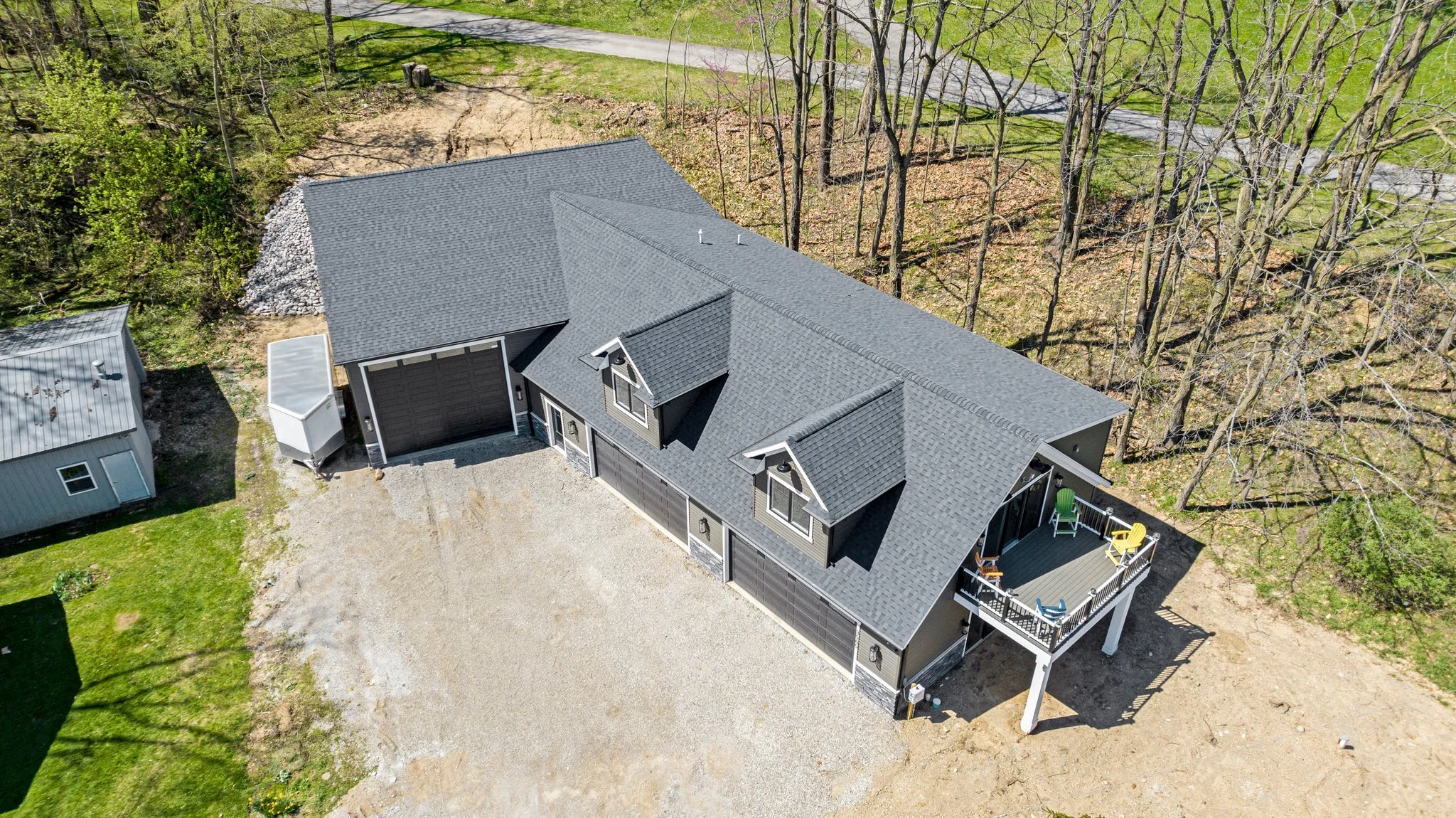 An aerial view of a modern house with a gray roof and black exterior walls, featuring a screened porch with outdoor chairs, surrounded by trees and a gravel driveway.