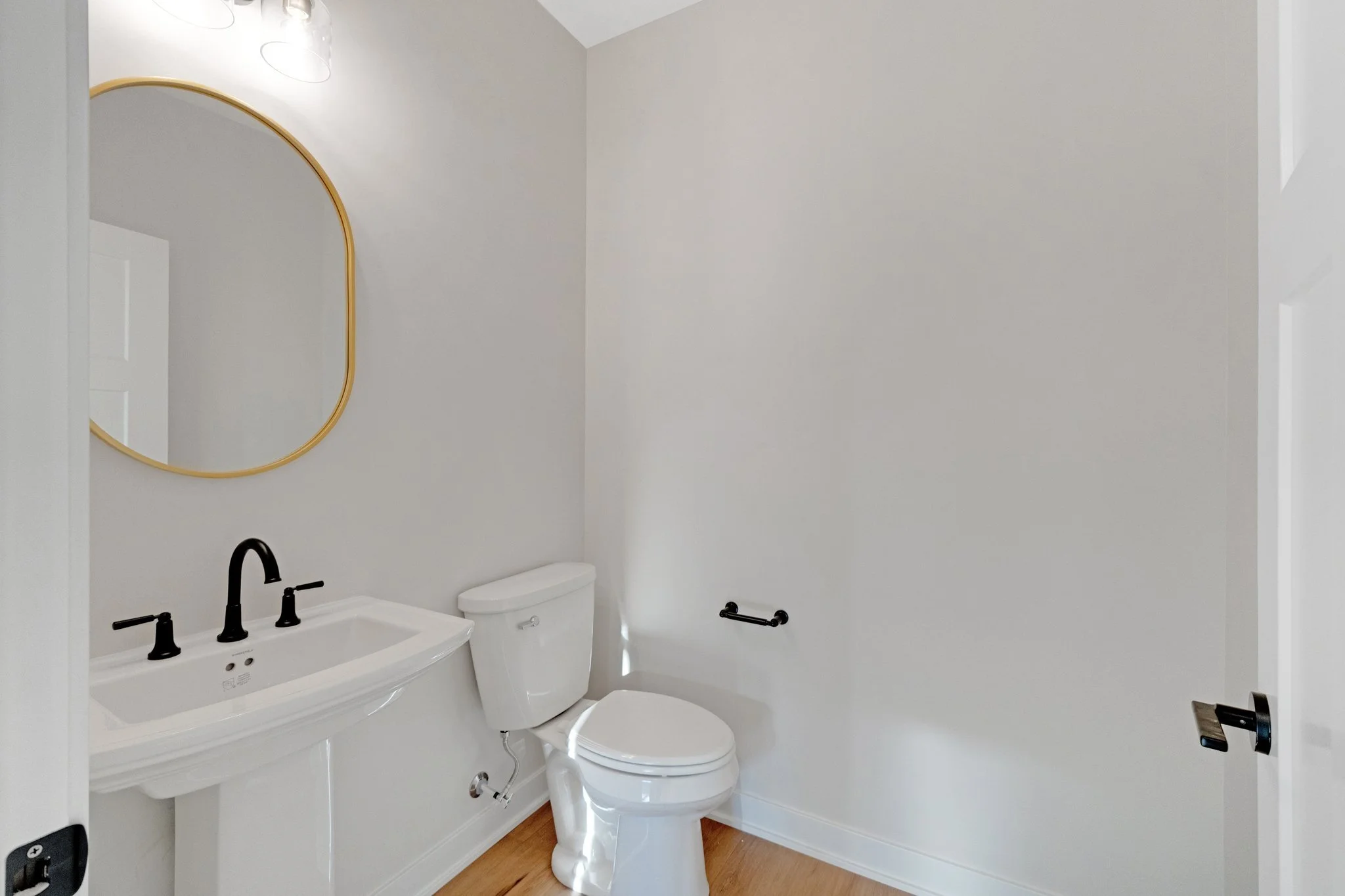 Small bathroom with a white toilet, a white sink with black faucet handles, a round mirror with a gold frame, and a wooden floor.