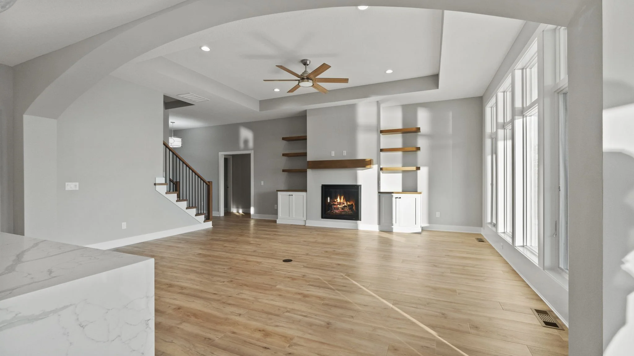 Empty living room with large windows, hardwood floors, a fireplace, built-in cabinets, and wooden shelves on the wall.