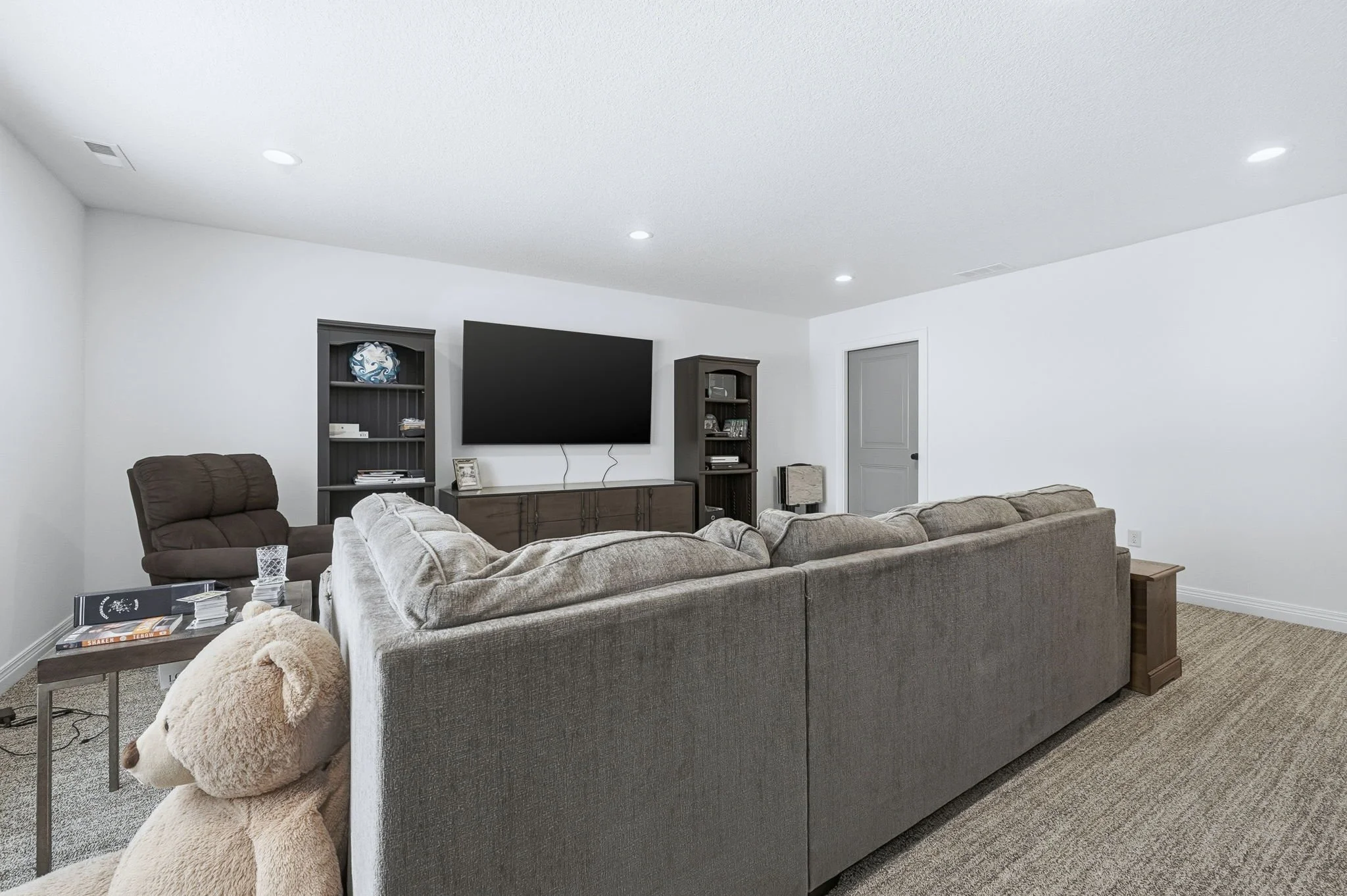 Living room with beige sectional couch, brown recliner, dark wood shelving unit with TV, white walls, gray door, and plush teddy bear on the carpeted floor.