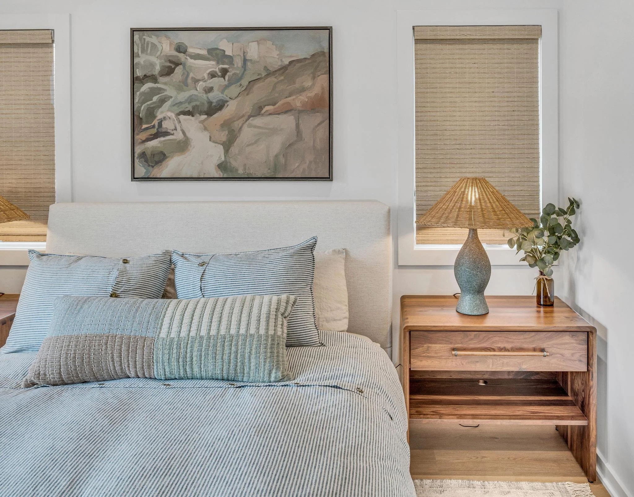 A bedroom with a white bed, striped pillows, a wooden nightstand, a lamp, a small plant in a vase, and a landscape painting on the wall.