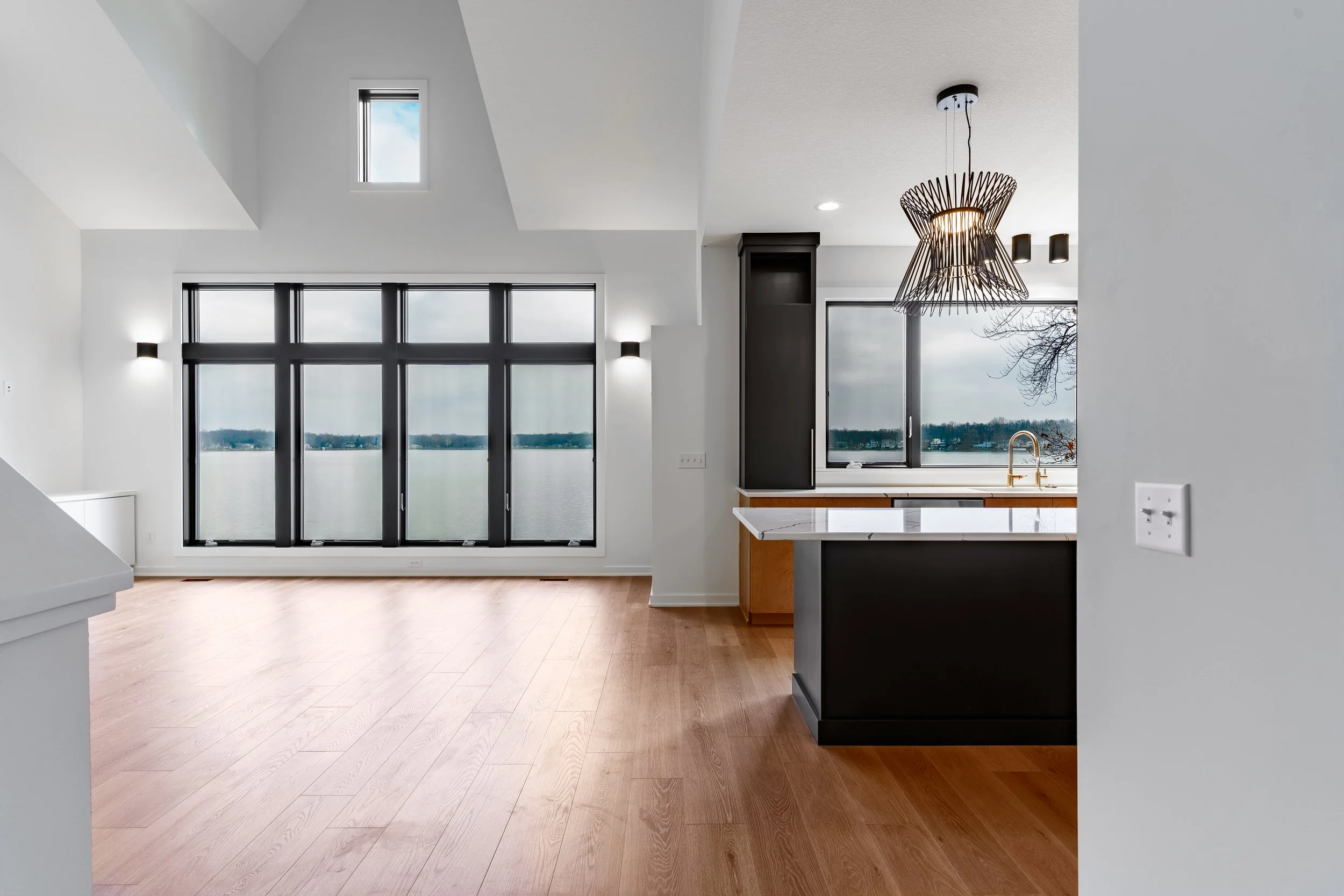 Modern kitchen and living area with large windows overlooking a lake, wooden flooring, black and white cabinets, and a pendant light.