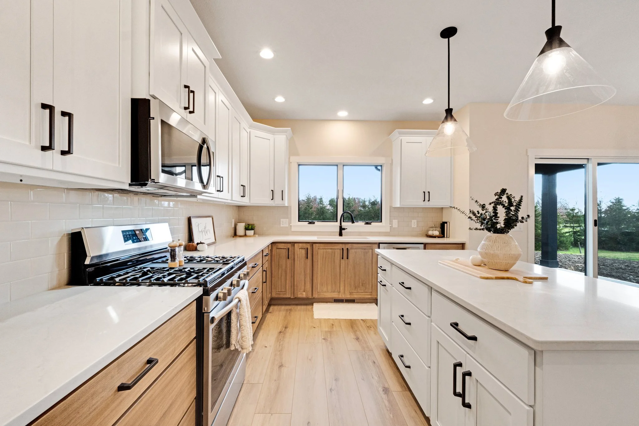 Modern kitchen with white upper cabinets, wood lower cabinets, white countertops, a window above the sink, stainless steel appliances, pendant lights, and a sliding glass door showing outdoor greenery.