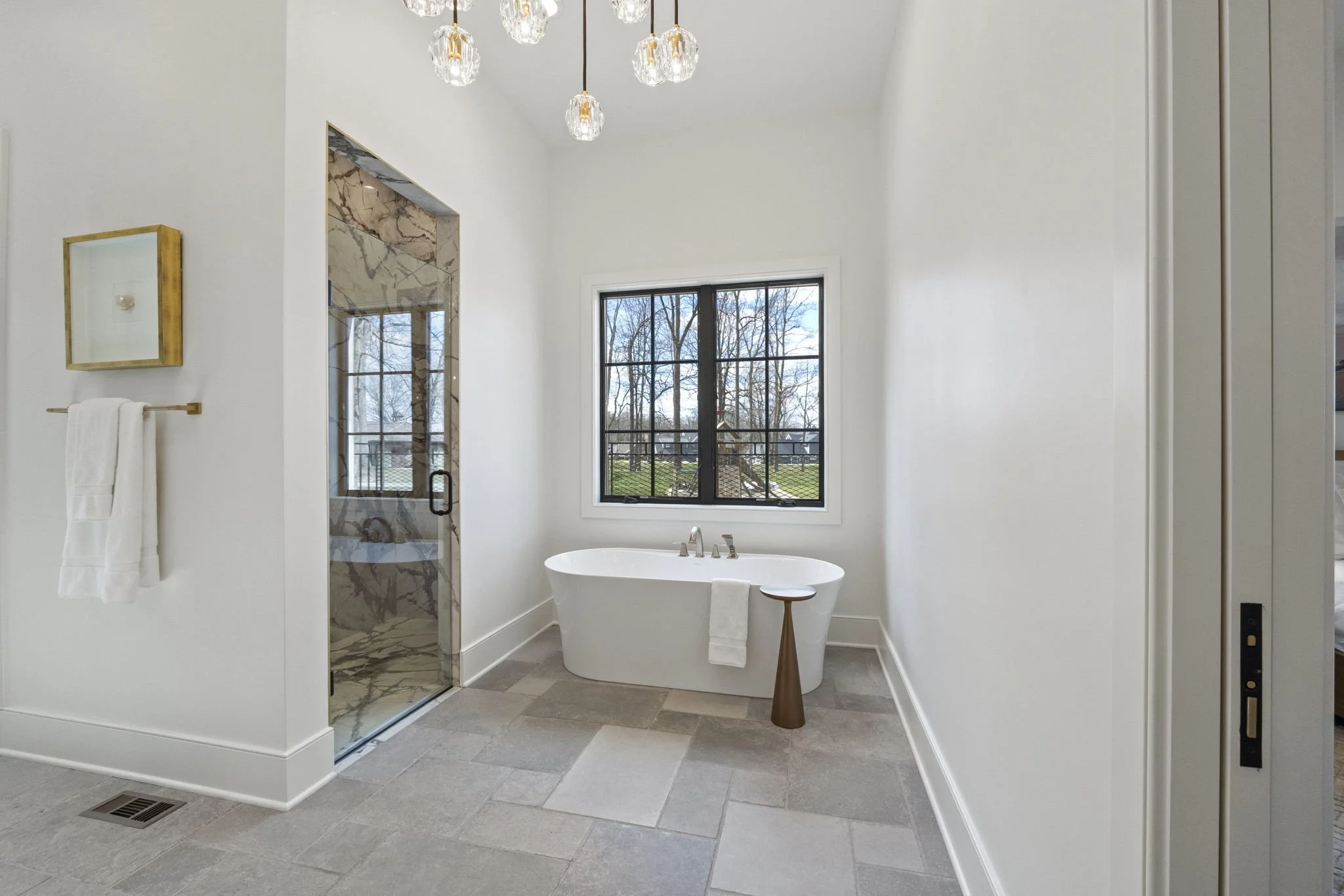Modern bathroom with a white bathtub, a window showing outdoor trees, a marble shower door, and minimal decor.