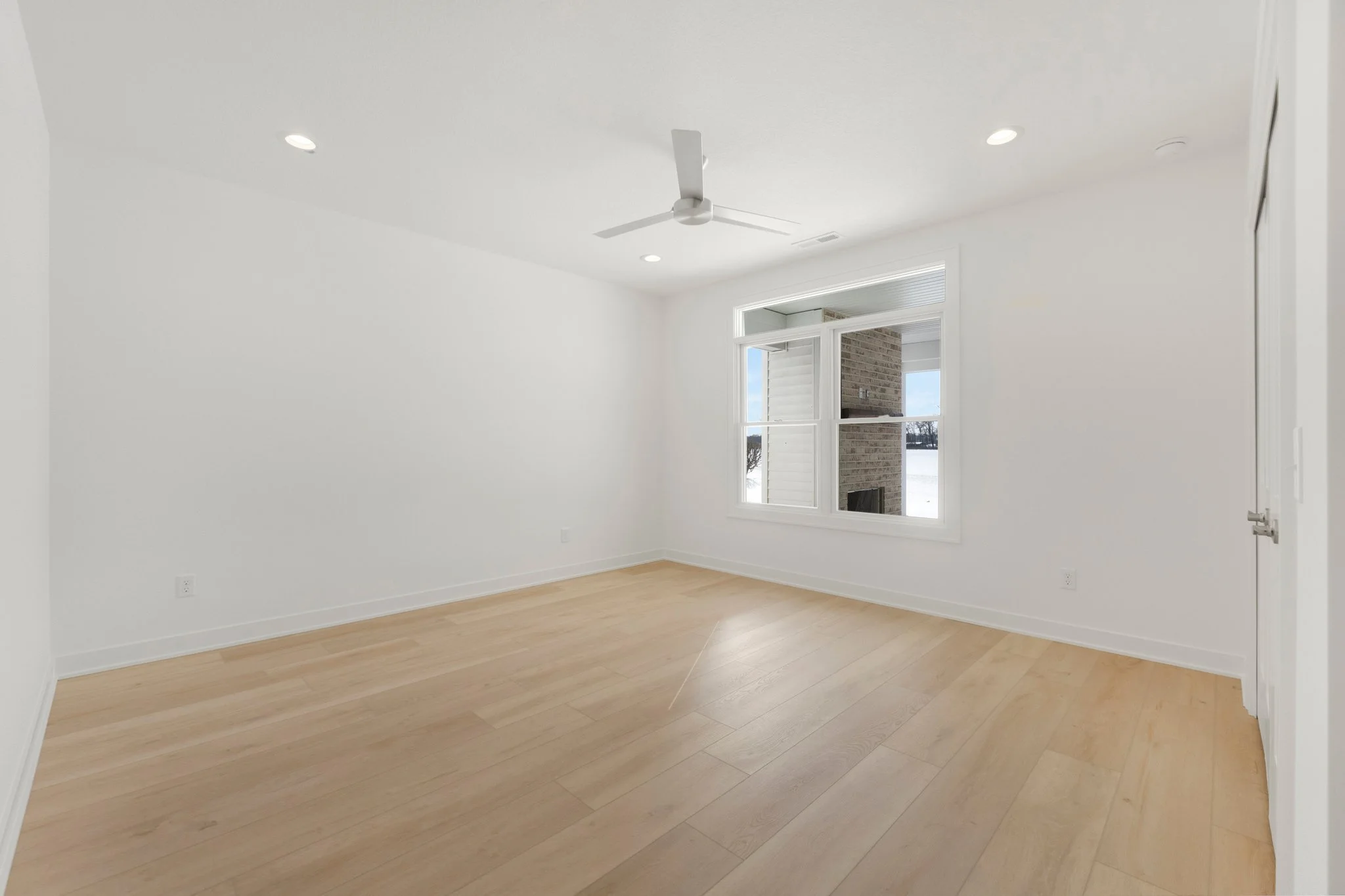 Empty room with white walls, light wood flooring, a ceiling fan, and a large window showing an outside view with snow and a brick chimney.