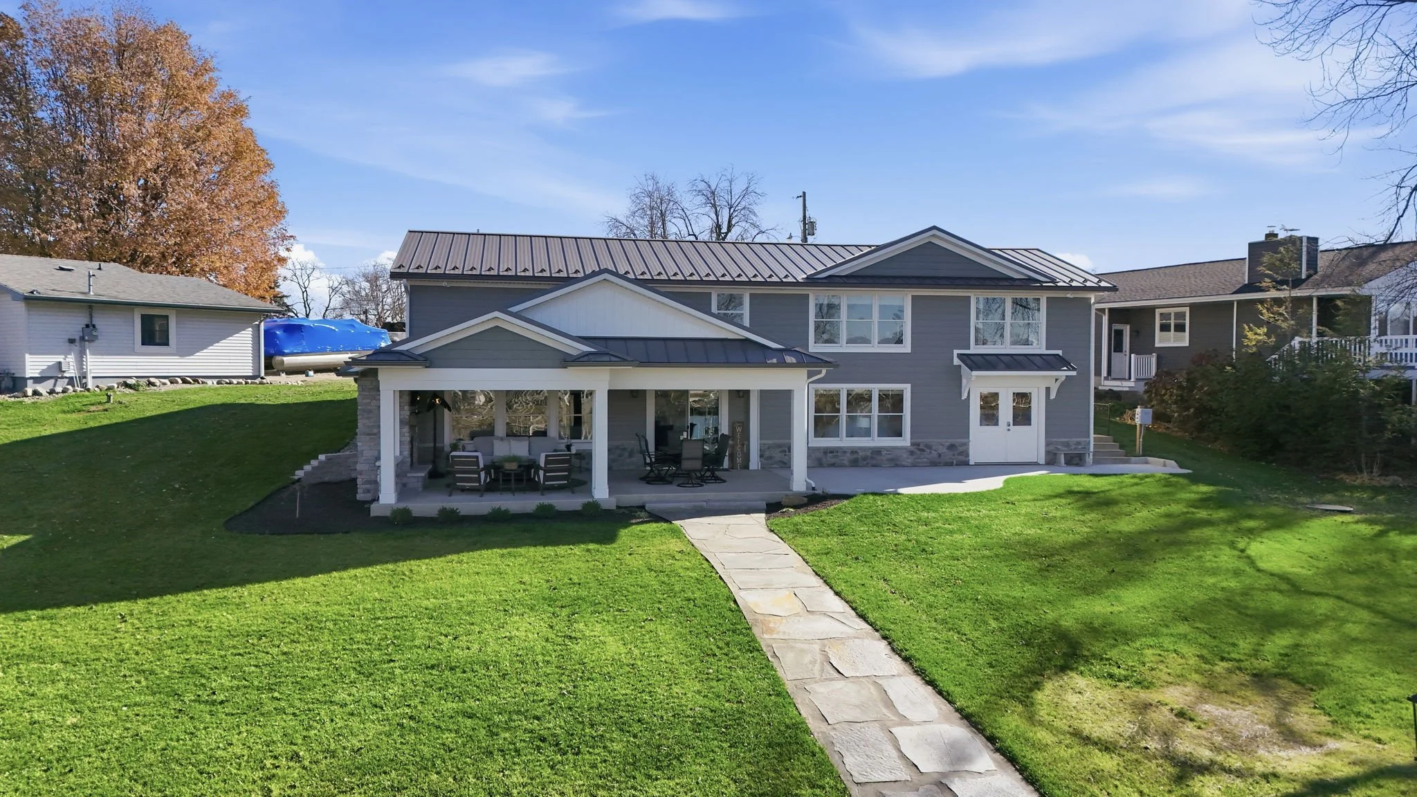 A two-story gray house with a metal roof, front porch with outdoor furniture, and a stone pathway leading to the yard. The lawn is green with some patches, and neighboring houses are visible on both sides. Trees with fall foliage are in the background.