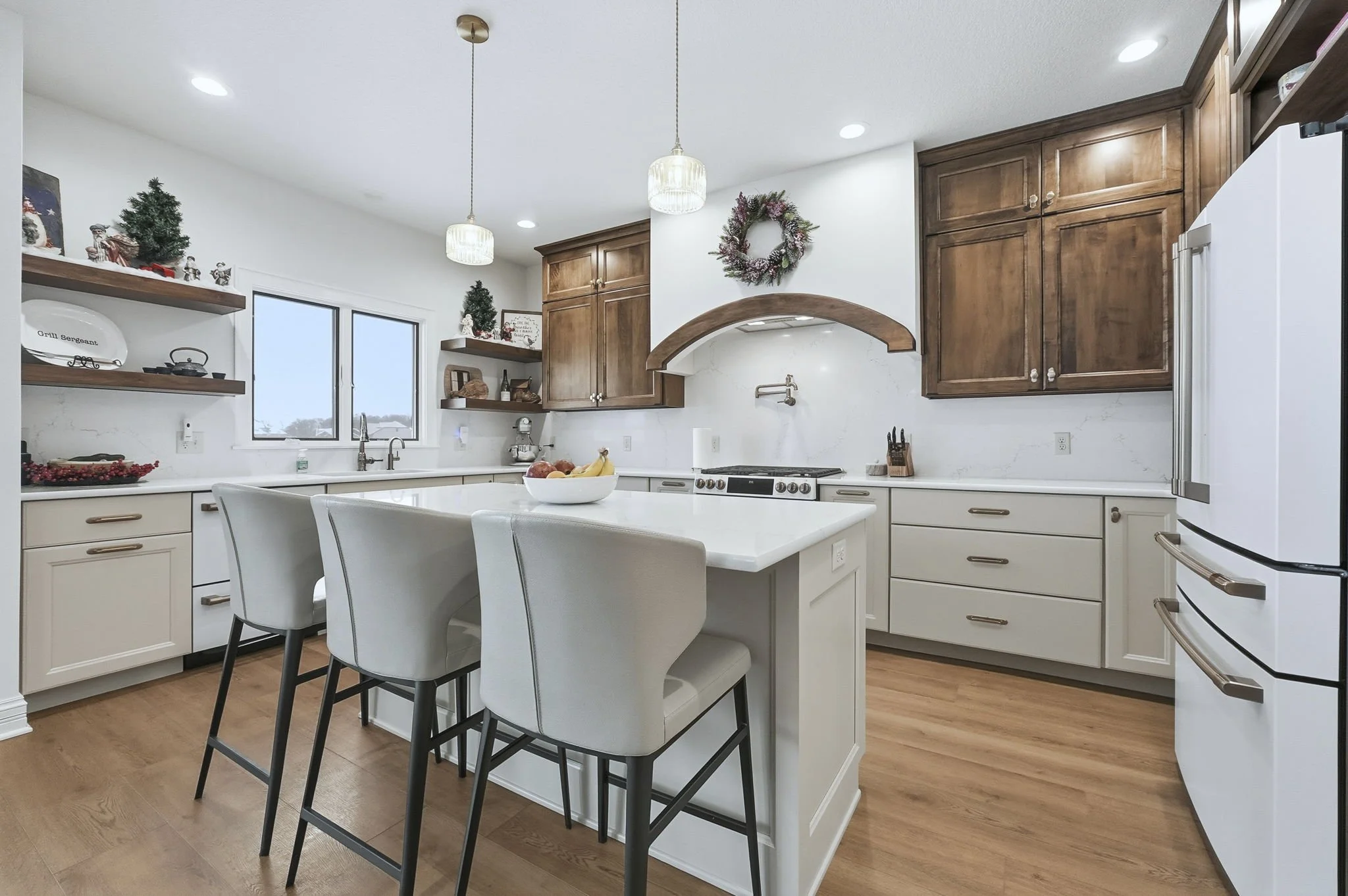 Modern kitchen with white counter, wooden cabinets, and a kitchen island with three bar stools. Decor includes festive Christmas ornaments, small trees, and a wreath on the wall.
