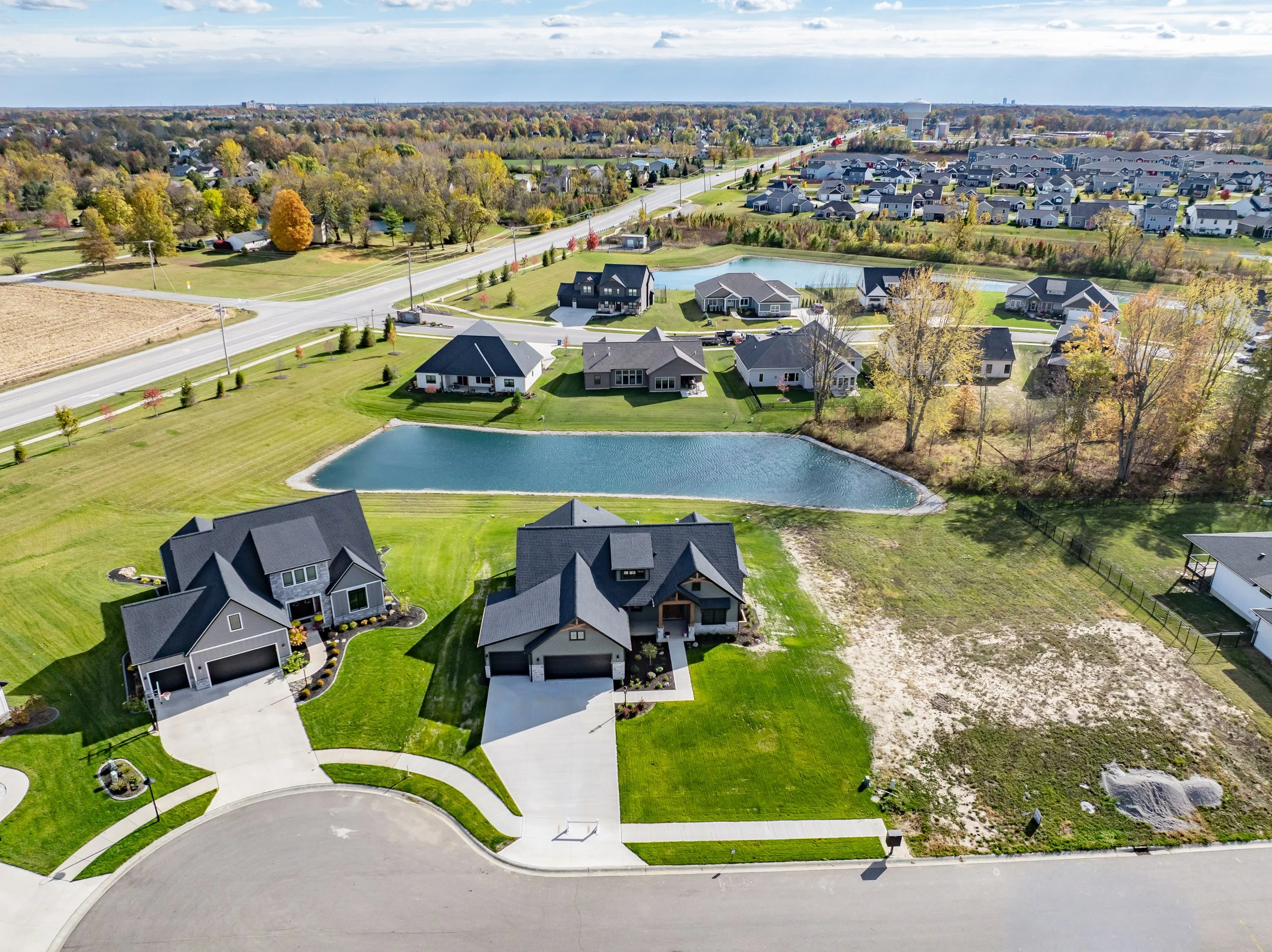 Aerial view of a suburban neighborhood with houses, green lawns, a pond, and trees, during daytime.