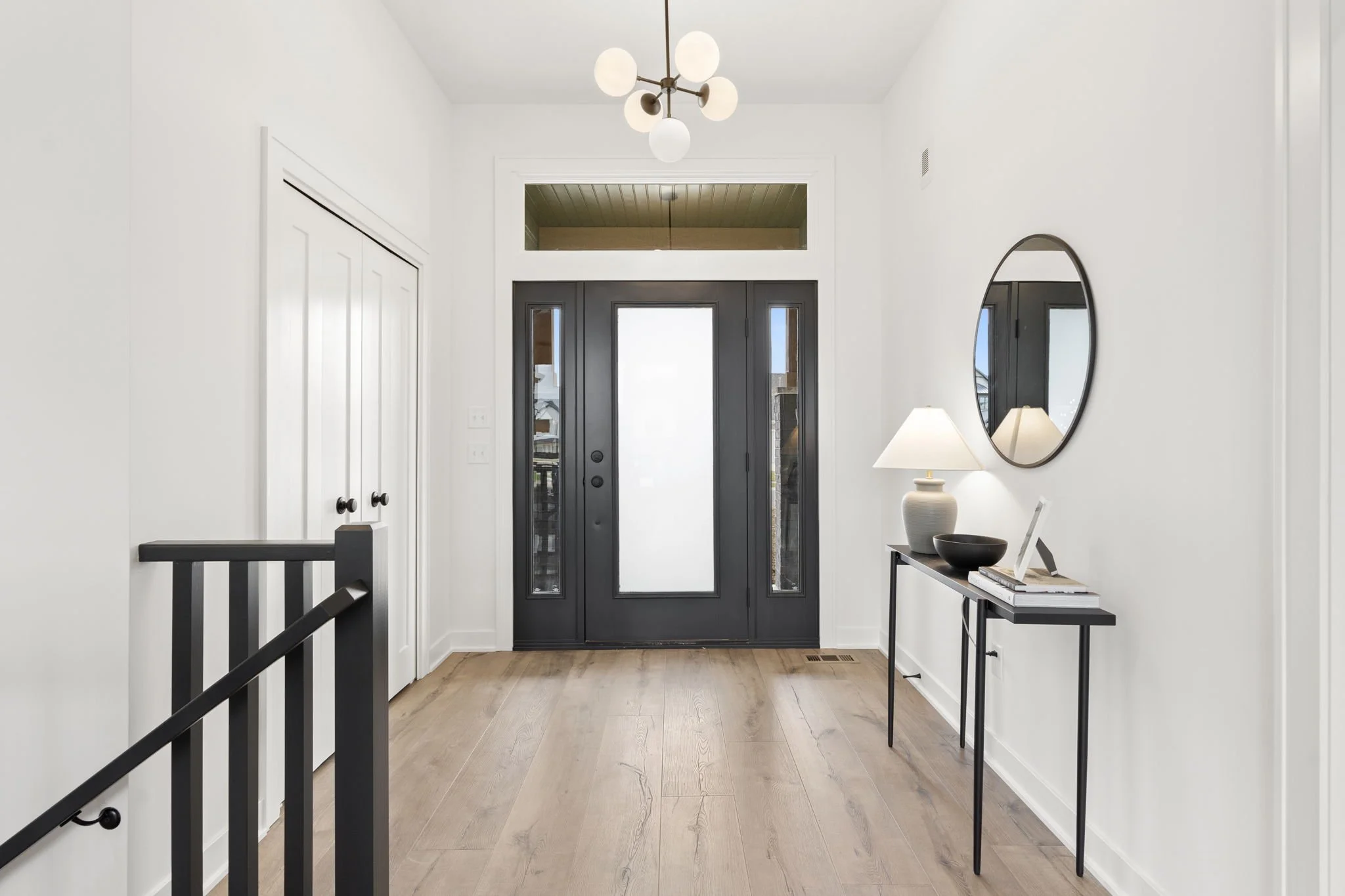 Modern entryway with a black front door, sidelight windows, wooden flooring, white walls, a mirror, a table with a lamp, bowl, and small books, and a chandelier.
