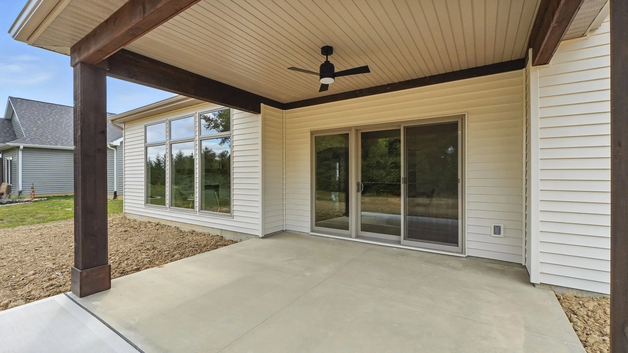 View of a newly built backyard patio with concrete flooring, white siding house with large windows, sliding glass door, and a covered porch with dark wooden support beams and a ceiling fan.