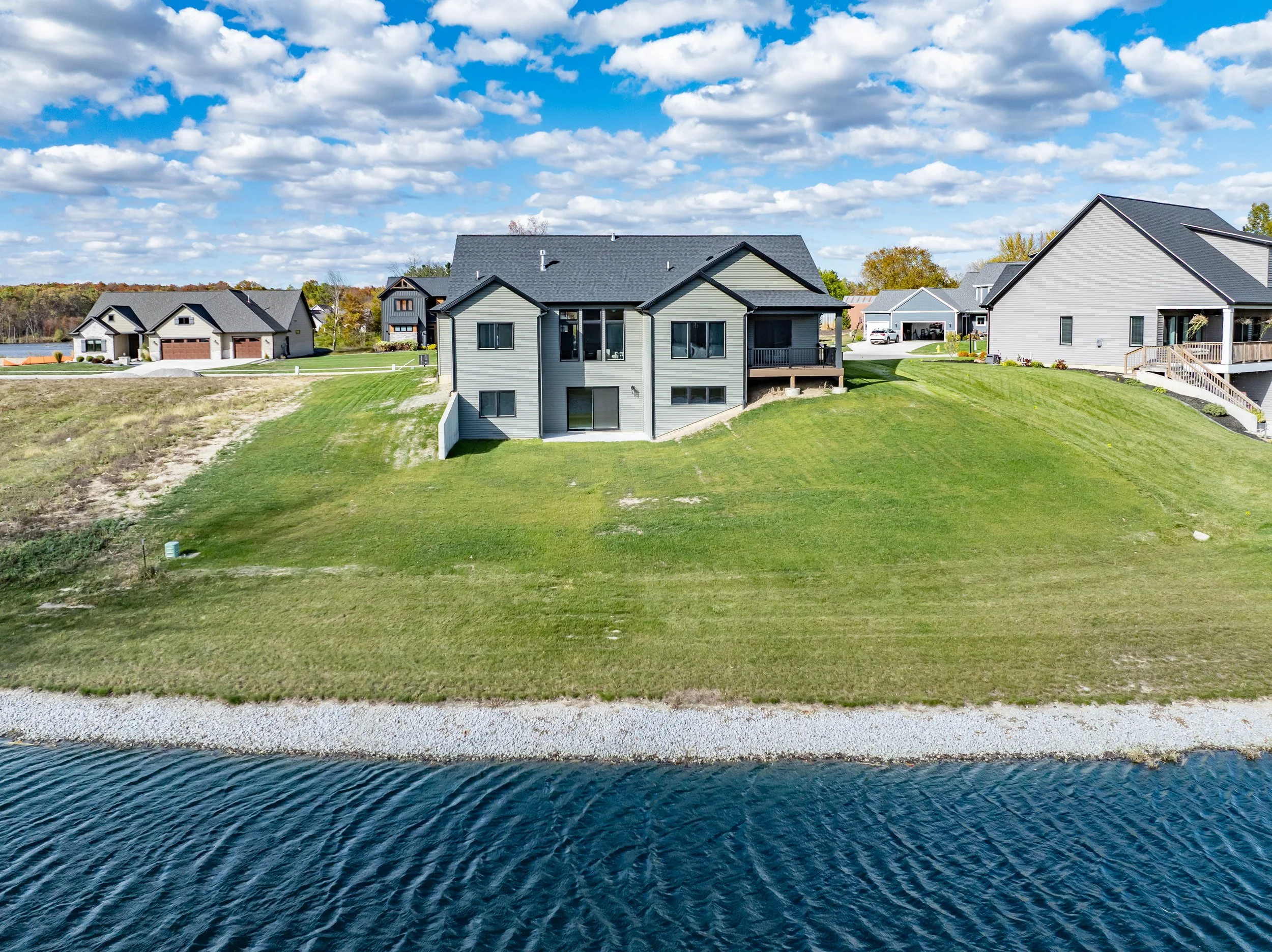 Aerial view of a modern house with gray siding, situated on a grassy hill beside a body of water, with other similar houses in the background under a partly cloudy sky.