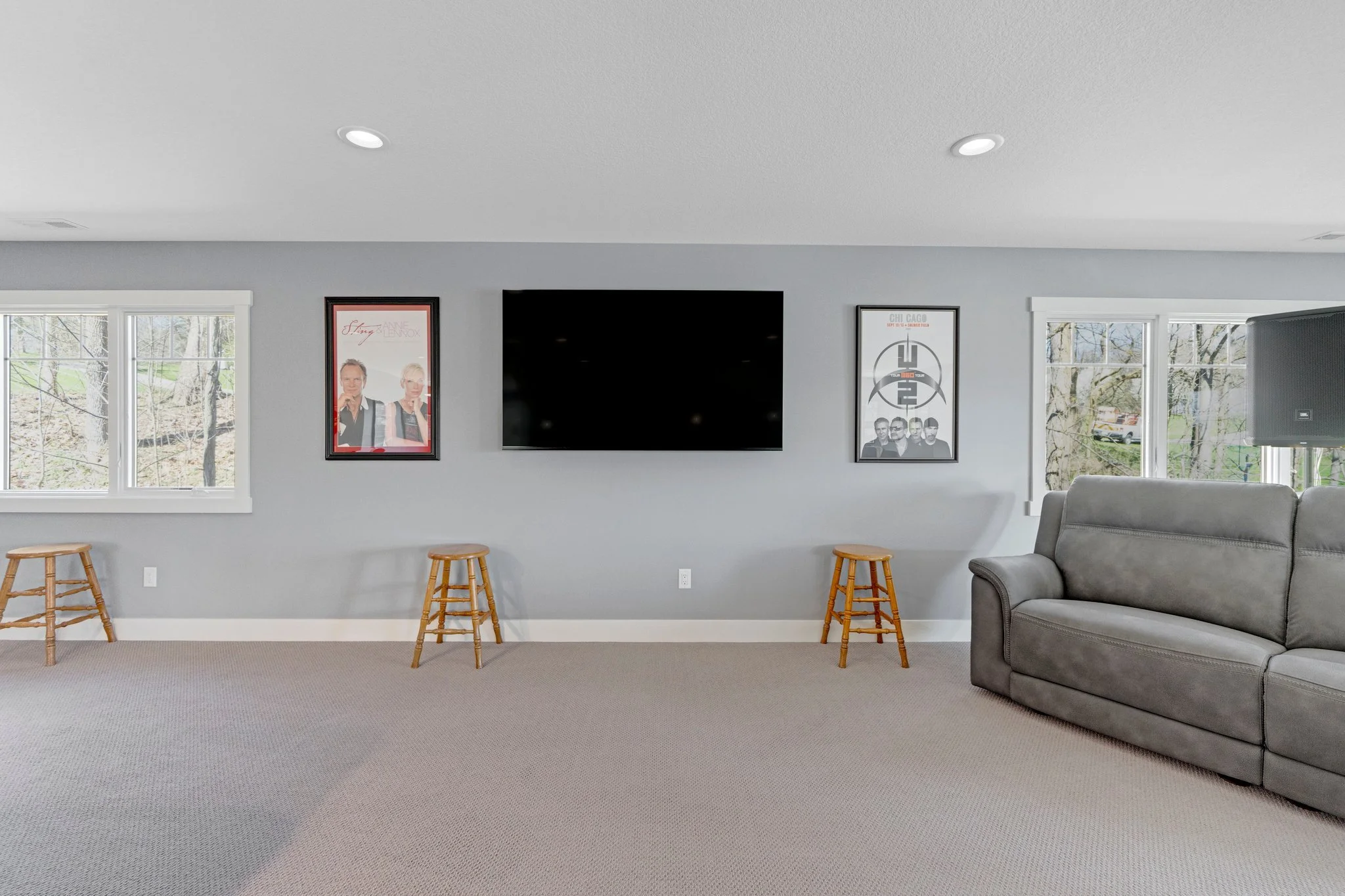 Living room with three posters on a light gray wall, a large flat-screen TV, and a gray sofa near windows showing an outdoor wooded area.