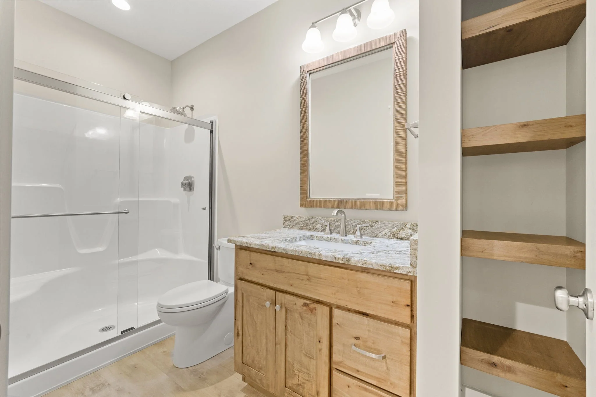 Bathroom with a glass shower enclosure, a white toilet, a wooden vanity with a granite countertop and a sink, a mirror with a wooden frame, and open wooden shelves on the wall.